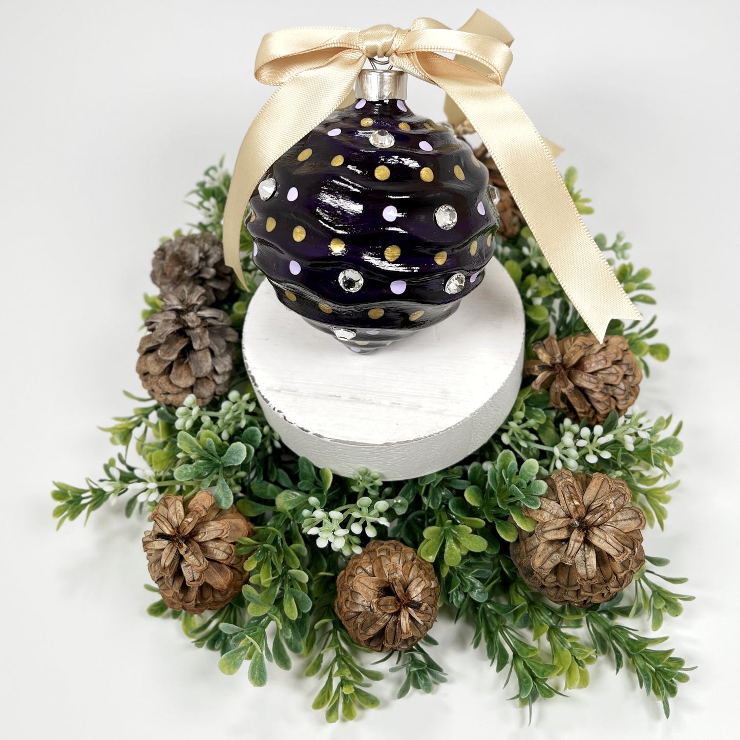 Decorative hand painted purple ornament with lavender and gold dots , rhinestones and a gold ribbon on a white base surrounded by greenery and pinecones.