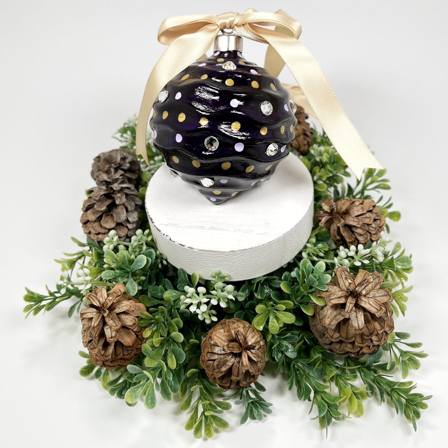 Decorative hand painted purple ornament with lavender and gold dots, rhinestones  and a gold ribbon on a white base surrounded by greenery and pinecones.