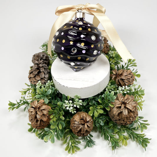 Decorative hand painted purple ornament with lavender and gold dots, rhinestones  and a gold ribbon on a white base surrounded by greenery and pinecones.
