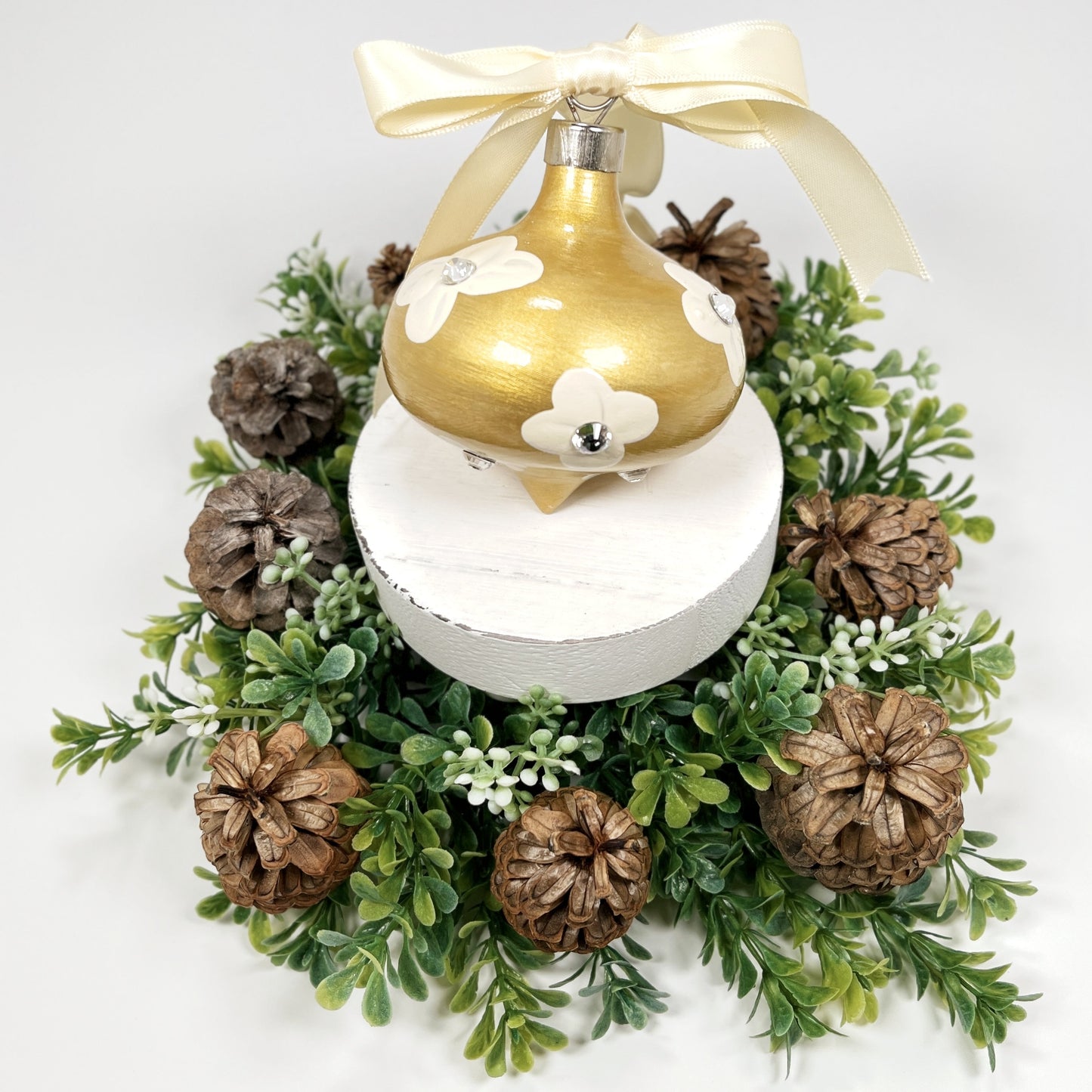 Decorative hand painted gold Christmas ornament with cream flowers with rhinestone centers and a cream bow on a white base with greenery and pine cones.