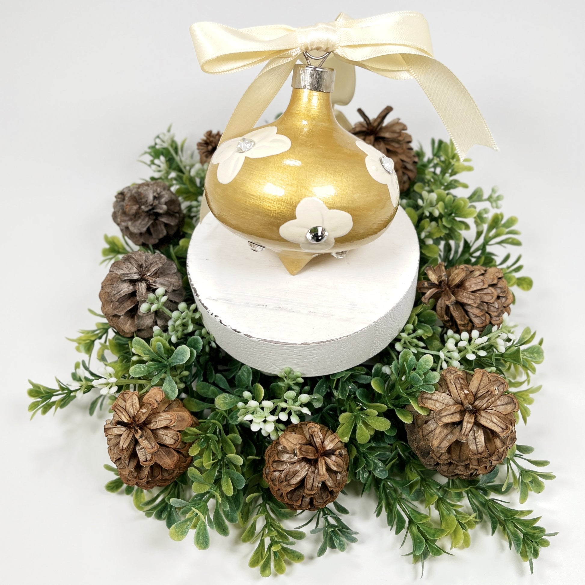 Decorative hand painted gold Christmas ornament with cream flowers with rhinestone centers and a cream bow on a white base with greenery and pine cones.