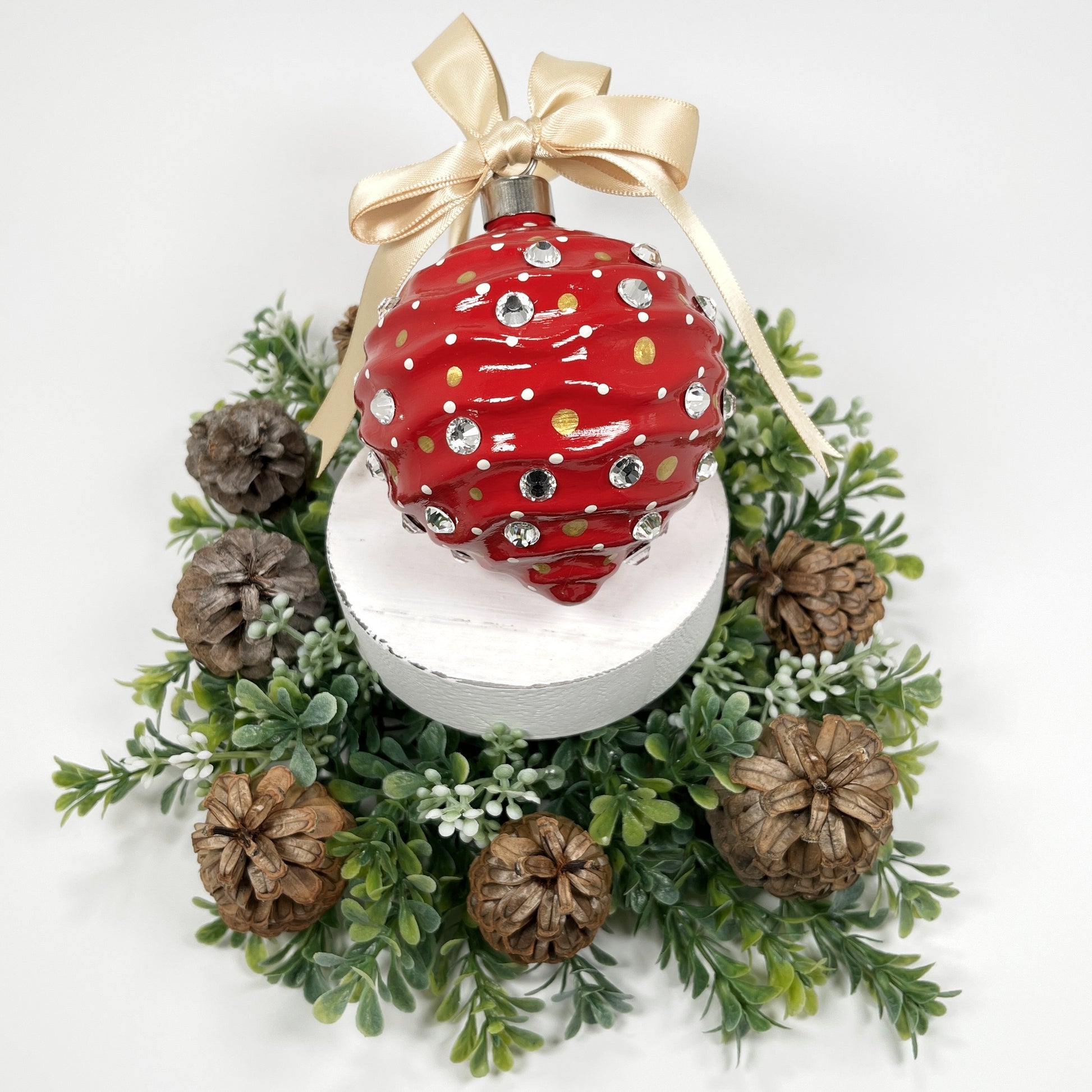 Decorative hand painted red Christmas ornament with gold and cream polka dots, rhinestone and a gold bow on a white base with greenery and pine cones.