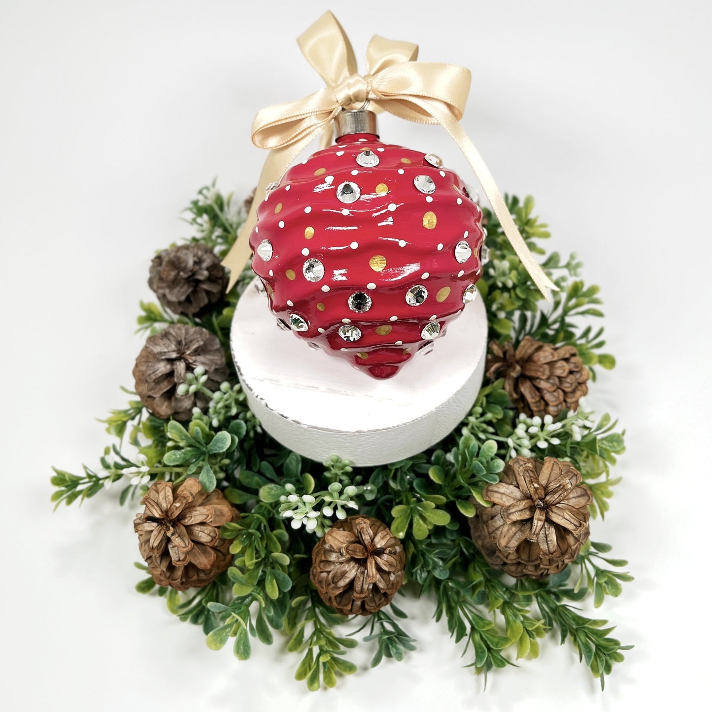 Decorative hand painted red Christmas ornament with gold and cream polka dots, rhinestone and a gold bow on a white base with greenery and pine cones.