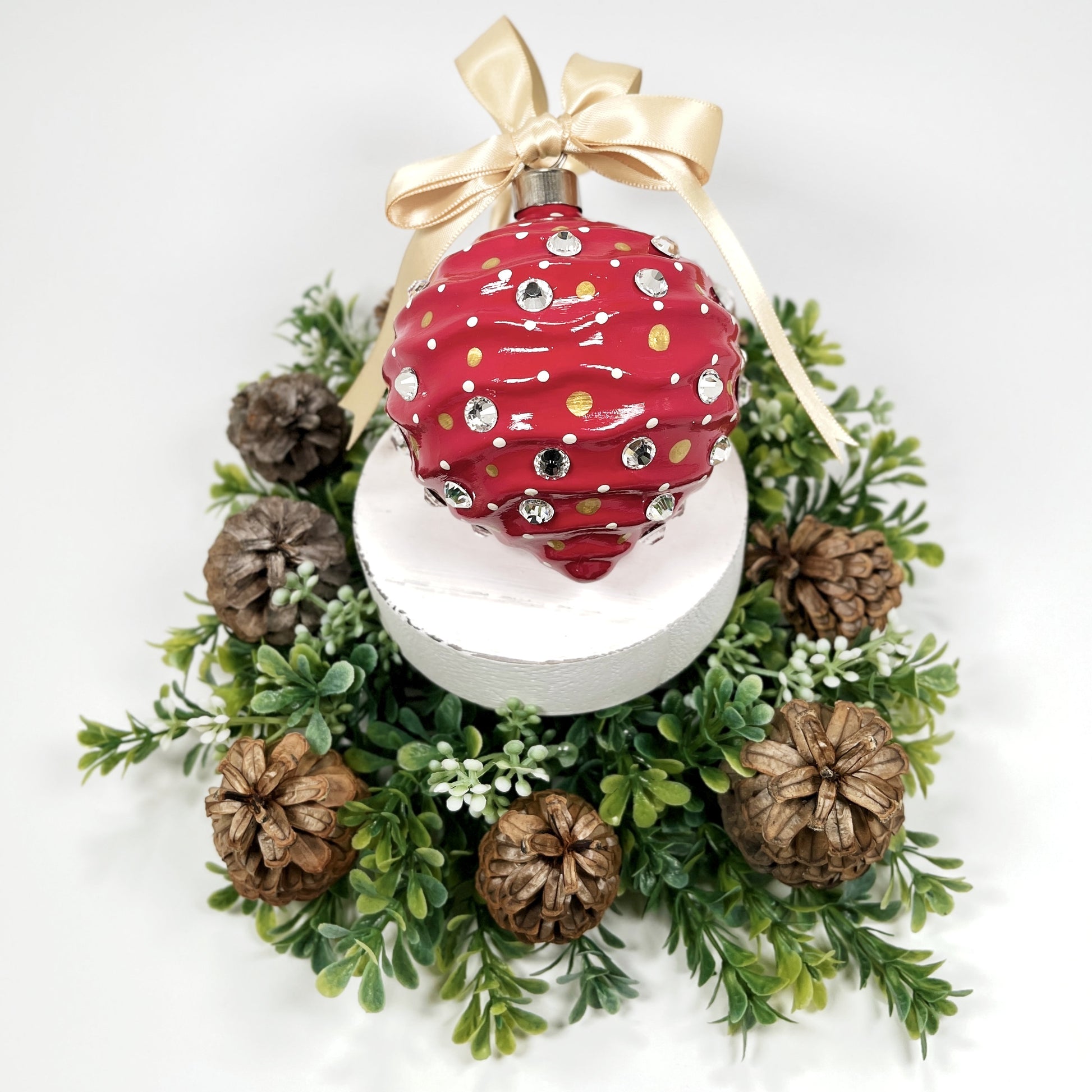 Decorative hand painted red Christmas ornament with gold and cream polka dots, rhinestone and a gold bow on a white base with greenery and pine cones.