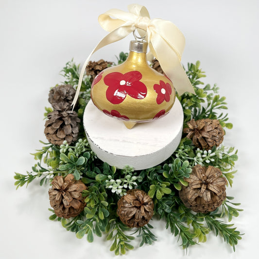 Decorative hand painted gold Christmas ornament with red flowers with a cream bow on a white base with greenery and pine cones.