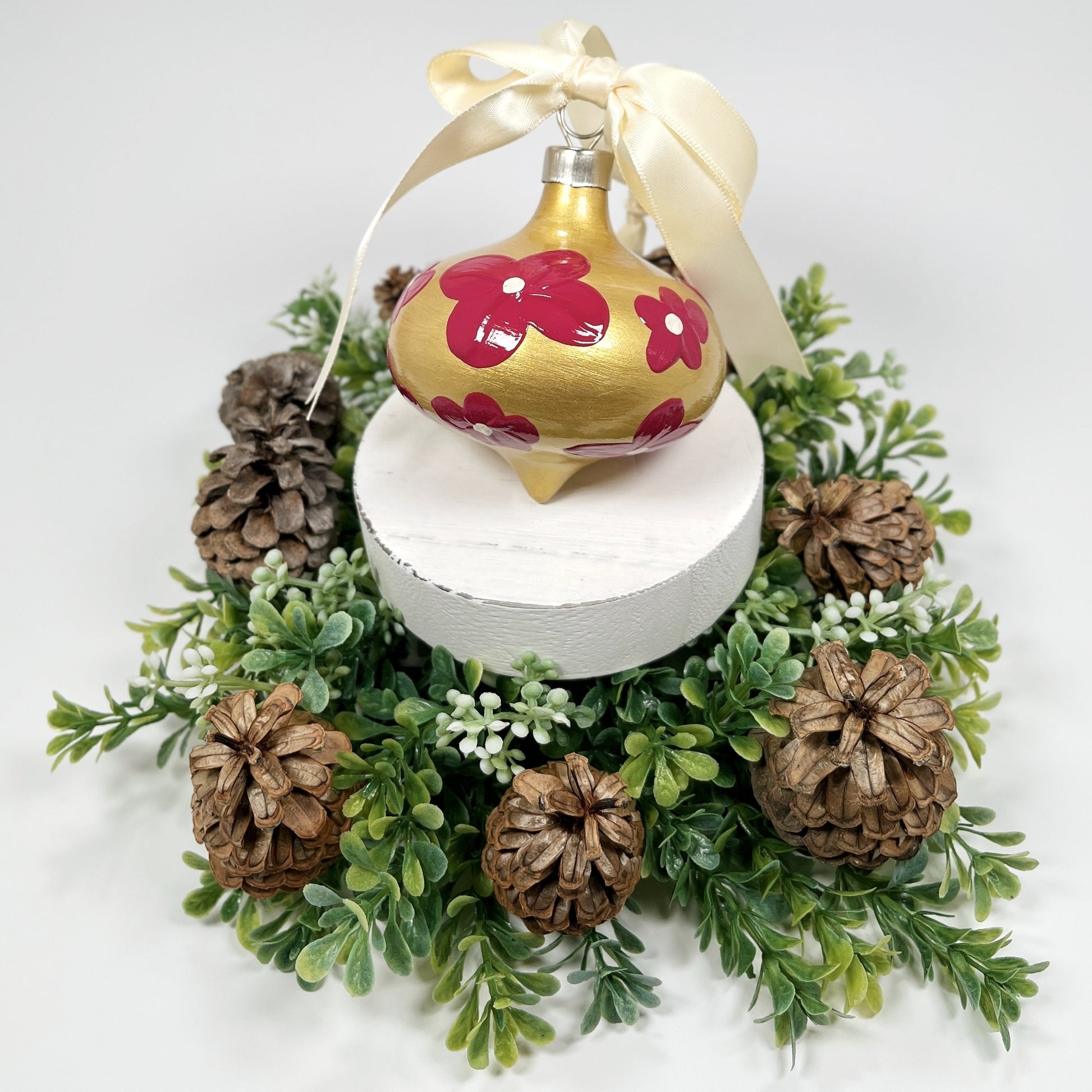 Decorative hand painted gold Christmas ornament with red flowers with a cream bow on a white base with greenery and pine cones.