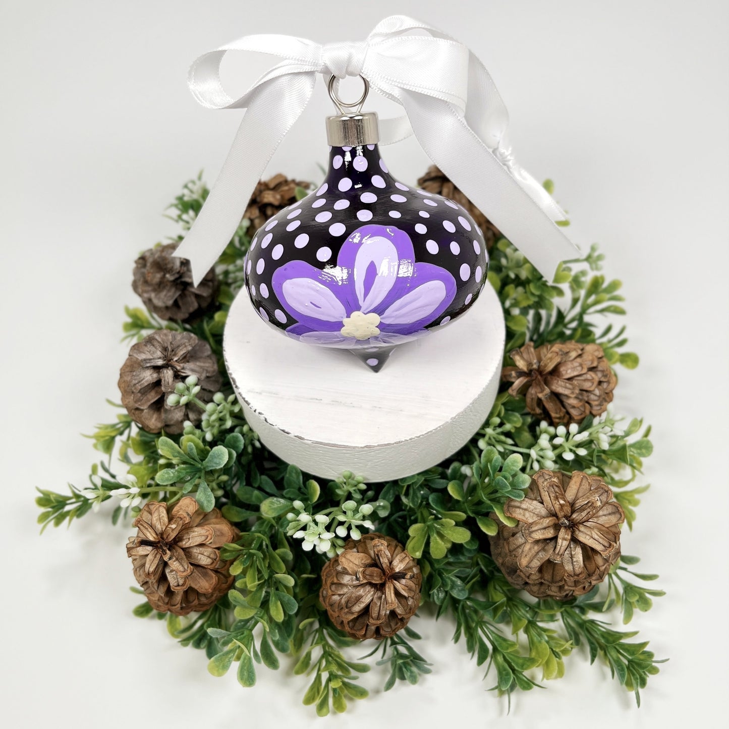 Decorative hand painted Christmas ornament with purple flowers and a white bow on a white base with greenery and pine cones.