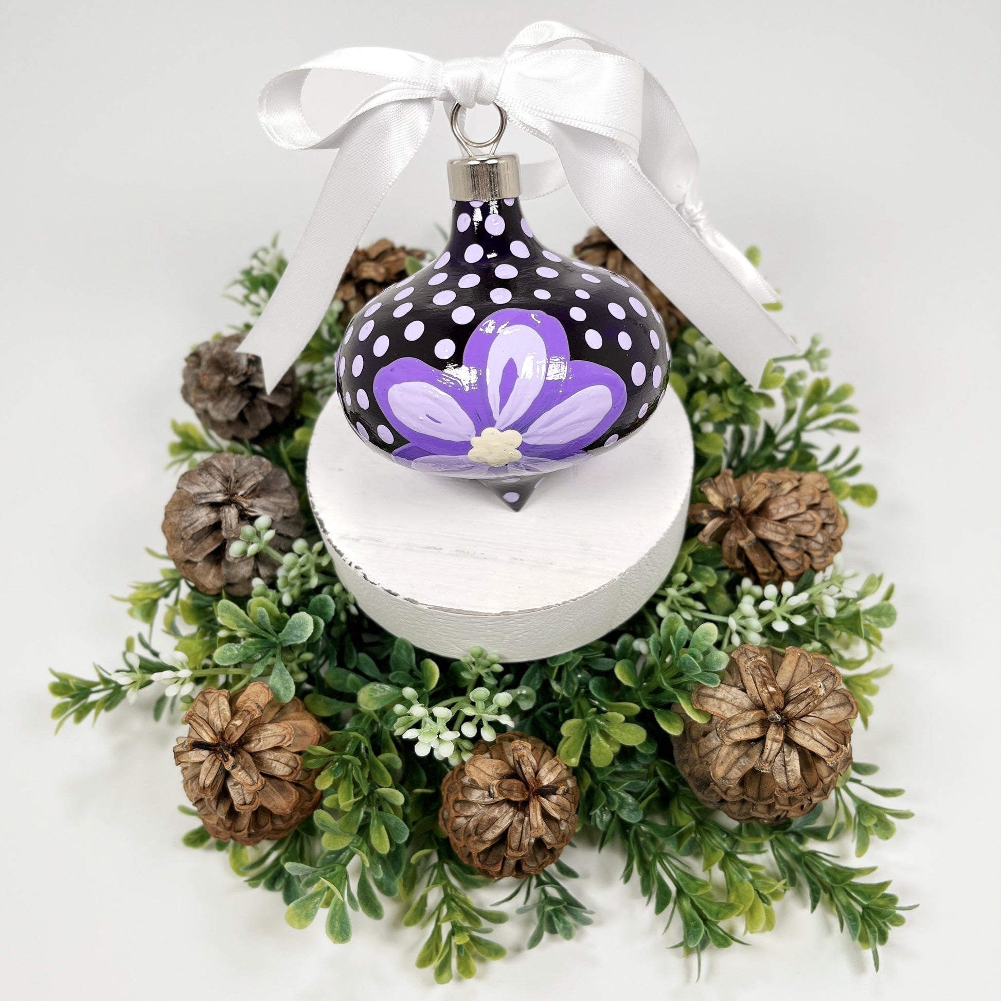 Decorative hand painted Christmas ornament with purple flowers and a white bow on a white base with greenery and pine cones.