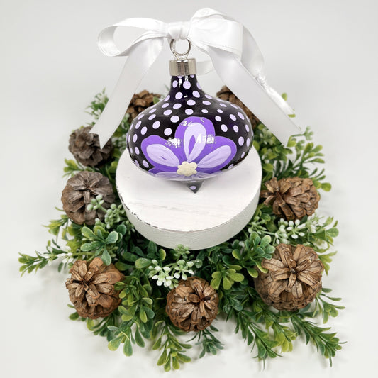 Decorative hand painted Christmas ornament with purple flowers and a white bow on a white base with greenery and pine cones.