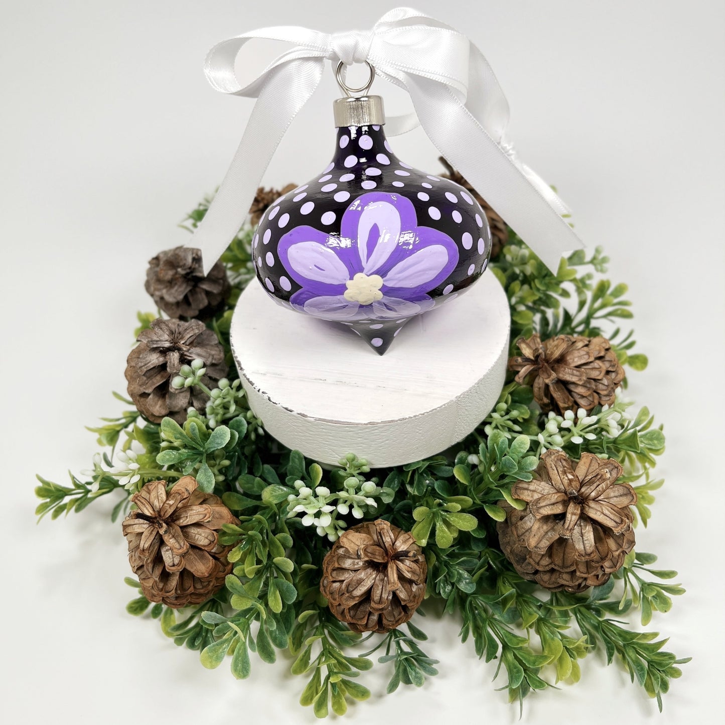 Decorative hand painted Christmas ornament with purple flowers and a white bow on a white base with greenery and pine cones.
