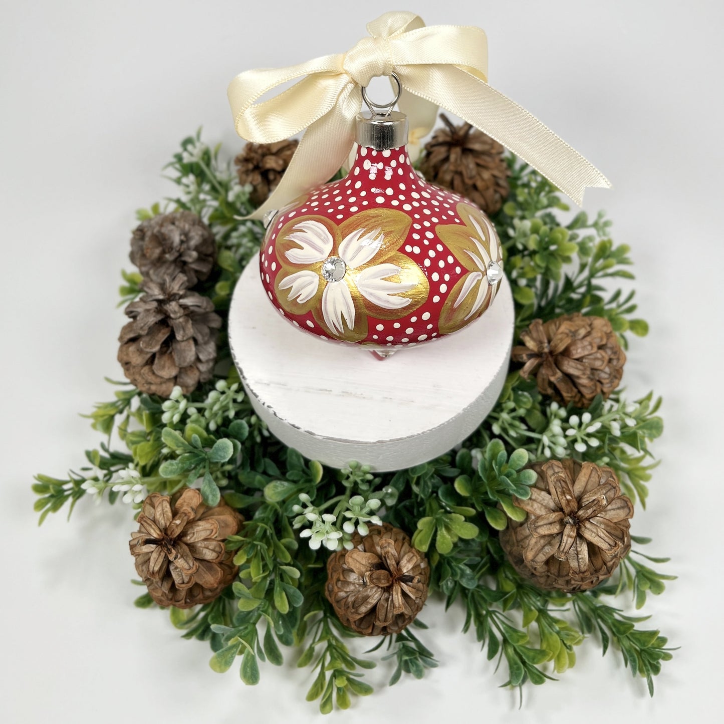 Decorative hand painted red Christmas ornament with gold and cream flowers with rhinestone centers, cream polka dots and a cream bow on a white base with greenery and pine cones.