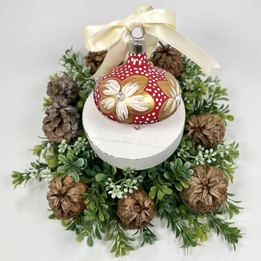 Decorative hand painted red Christmas ornament with gold and cream flowers with rhinestone centers, cream polka dots and a cream bow on a white base with greenery and pine cones.
