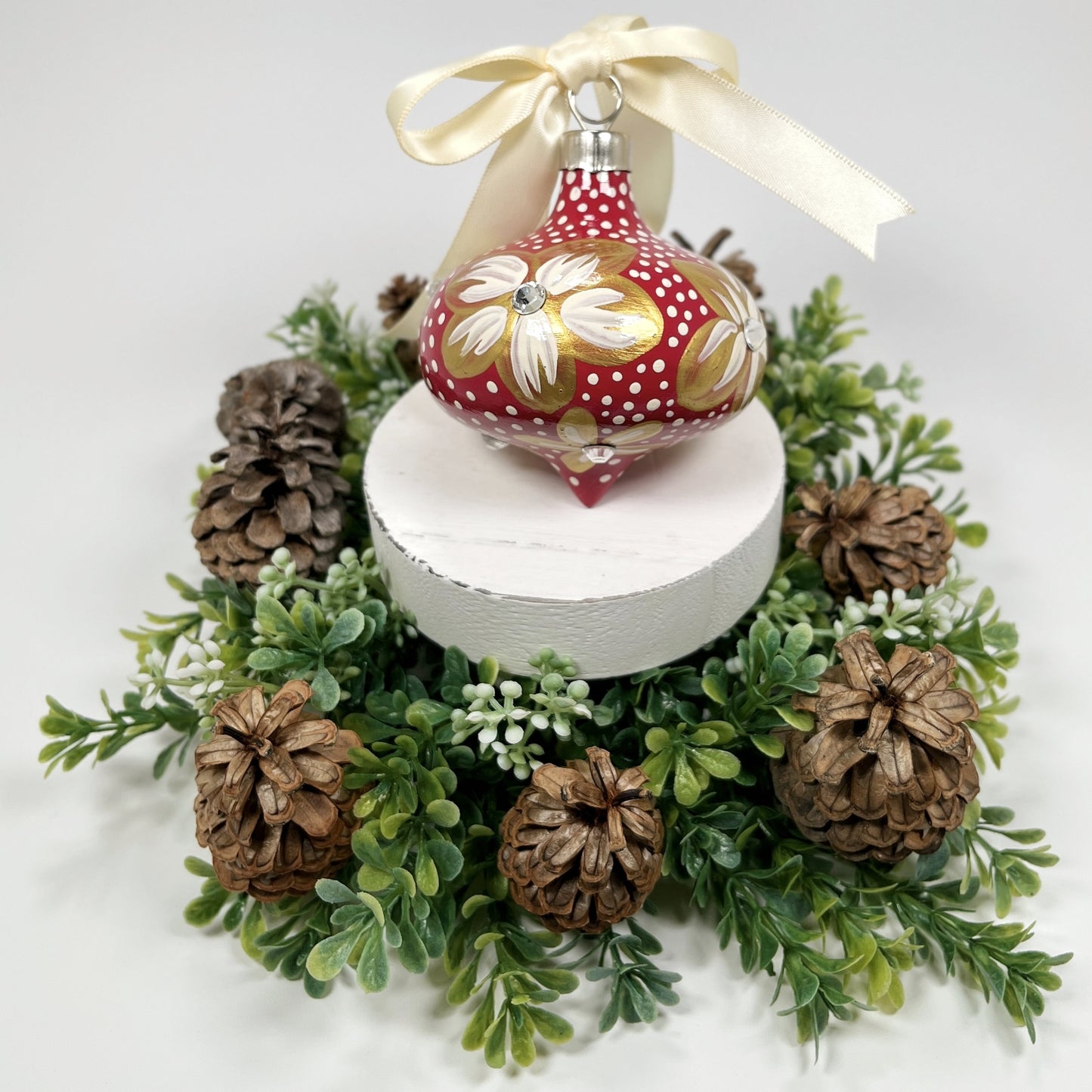 Decorative hand painted red Christmas ornament with gold and cream flowers with rhinestone centers, cream polka dots and a cream bow on a white base with greenery and pine cones.