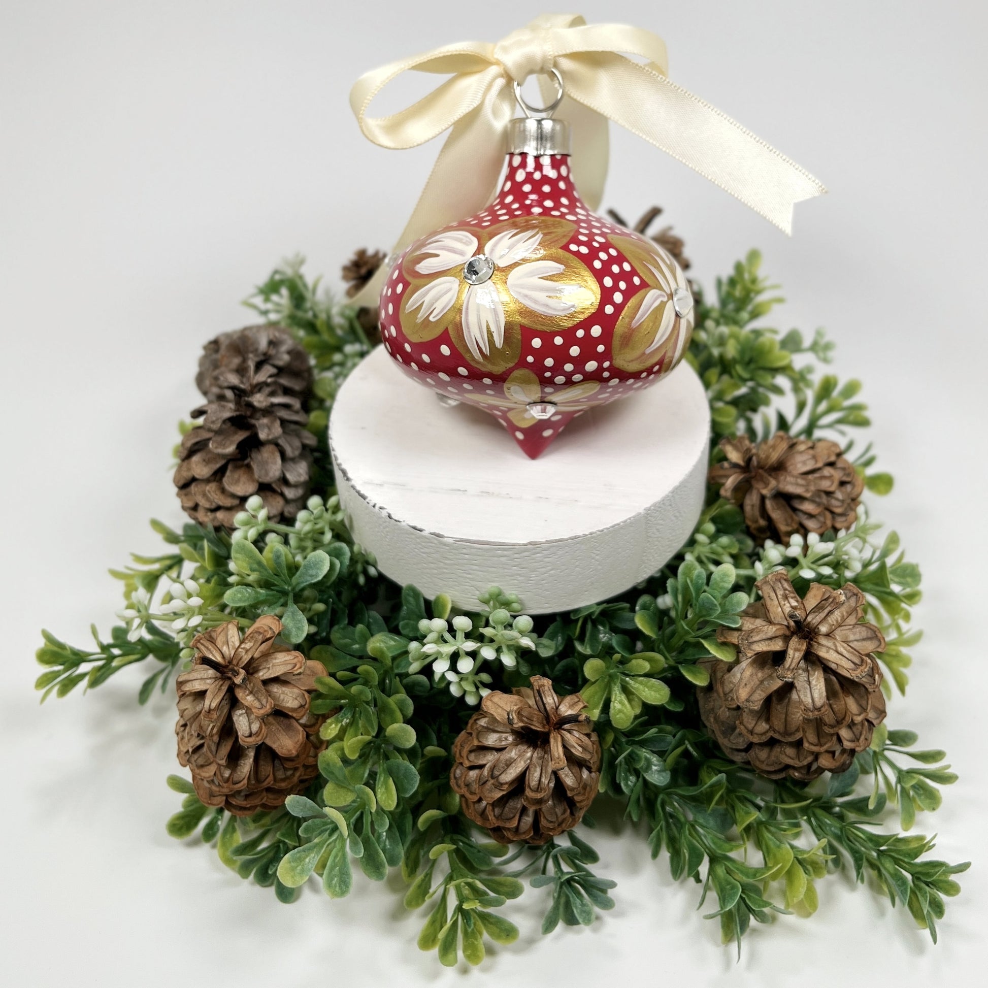 Decorative hand painted red Christmas ornament with gold and cream flowers with rhinestone centers, cream polka dots and a cream bow on a white base with greenery and pine cones.