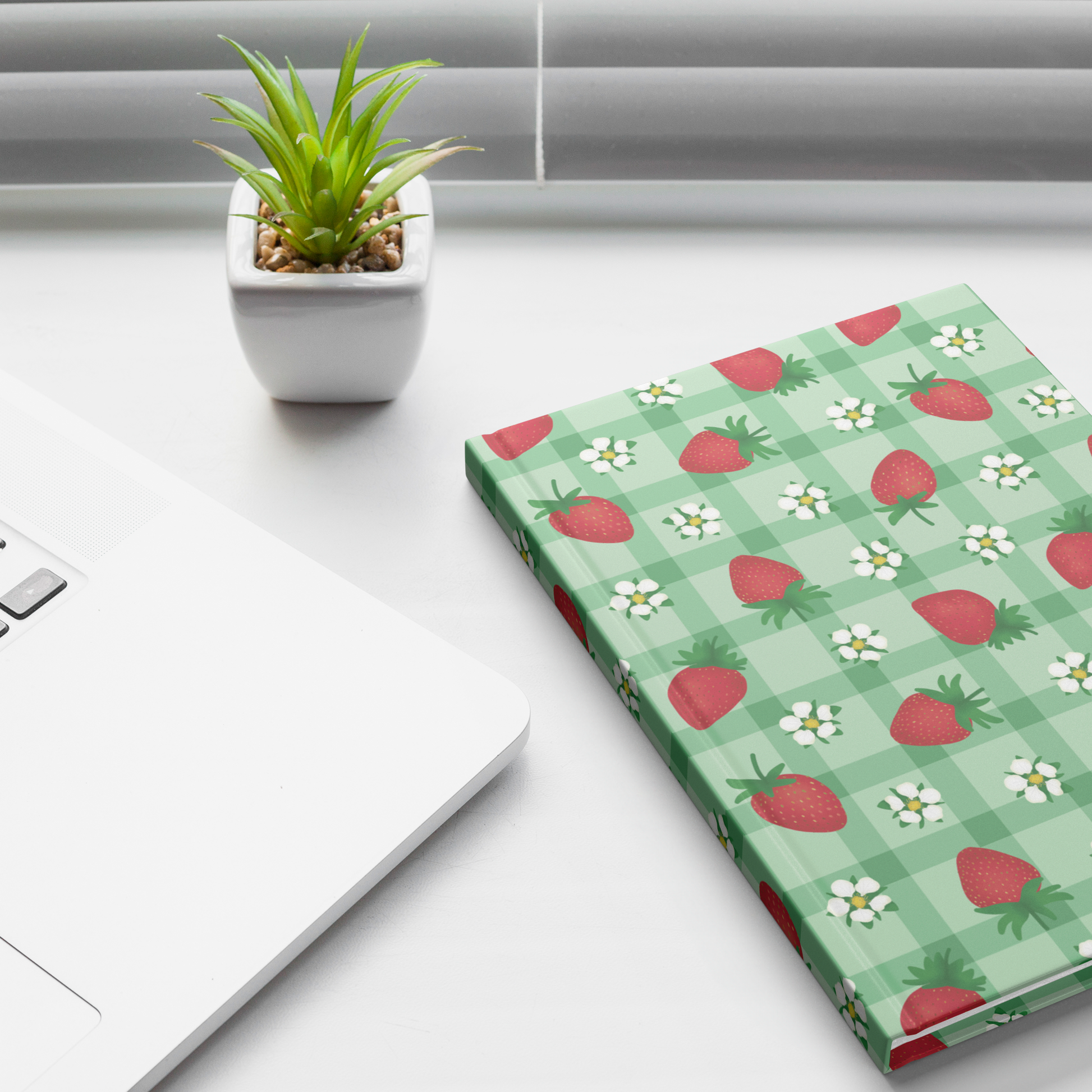 Hardcover journal with red strawberries and white flowers on green gingham sitting on a desk.