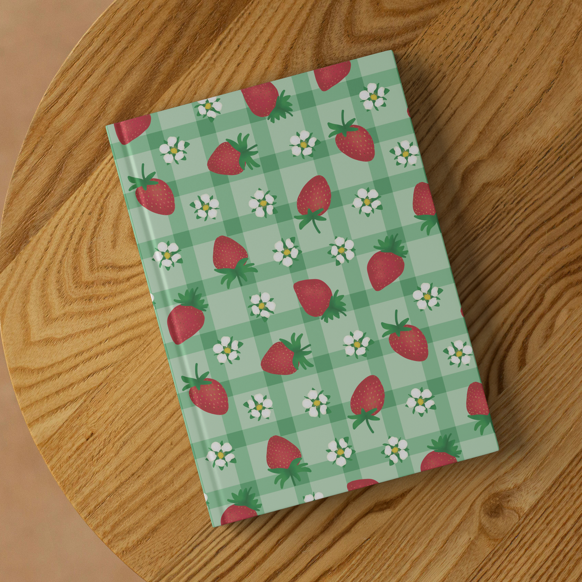 Hardcover journal with red strawberries and white flowers on green gingham, sitting on a wood table.