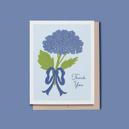 A greeting card with a white border, featuring a hand drawn illustration of blue hydrangea flowers with green leaves tied with a blue ribbon, next to the words 'Thank You' in blue script. The card is shown with a beige envelope behind it on a blue background.