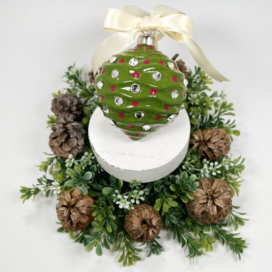 Decorative hand painted green Christmas ornament with fuchsia dots and rhinestones with a cream bow on a white base with greenery and pine cones.