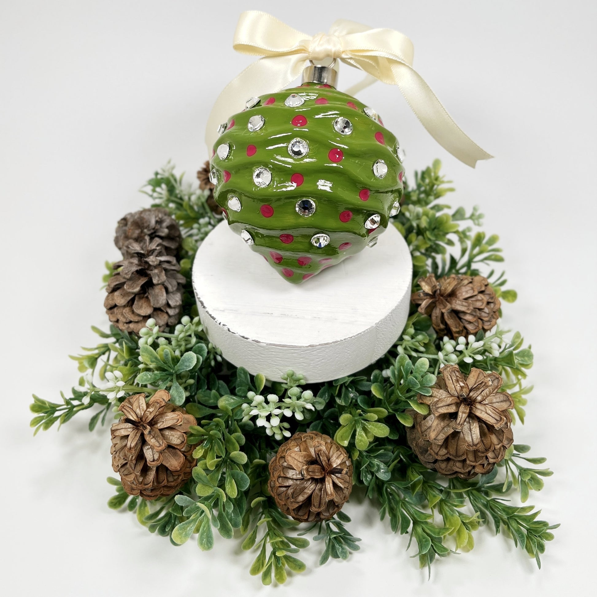 Decorative hand painted green Christmas ornament with fuchsia dots and rhinestones with a cream bow on a white base with greenery and pine cones.
