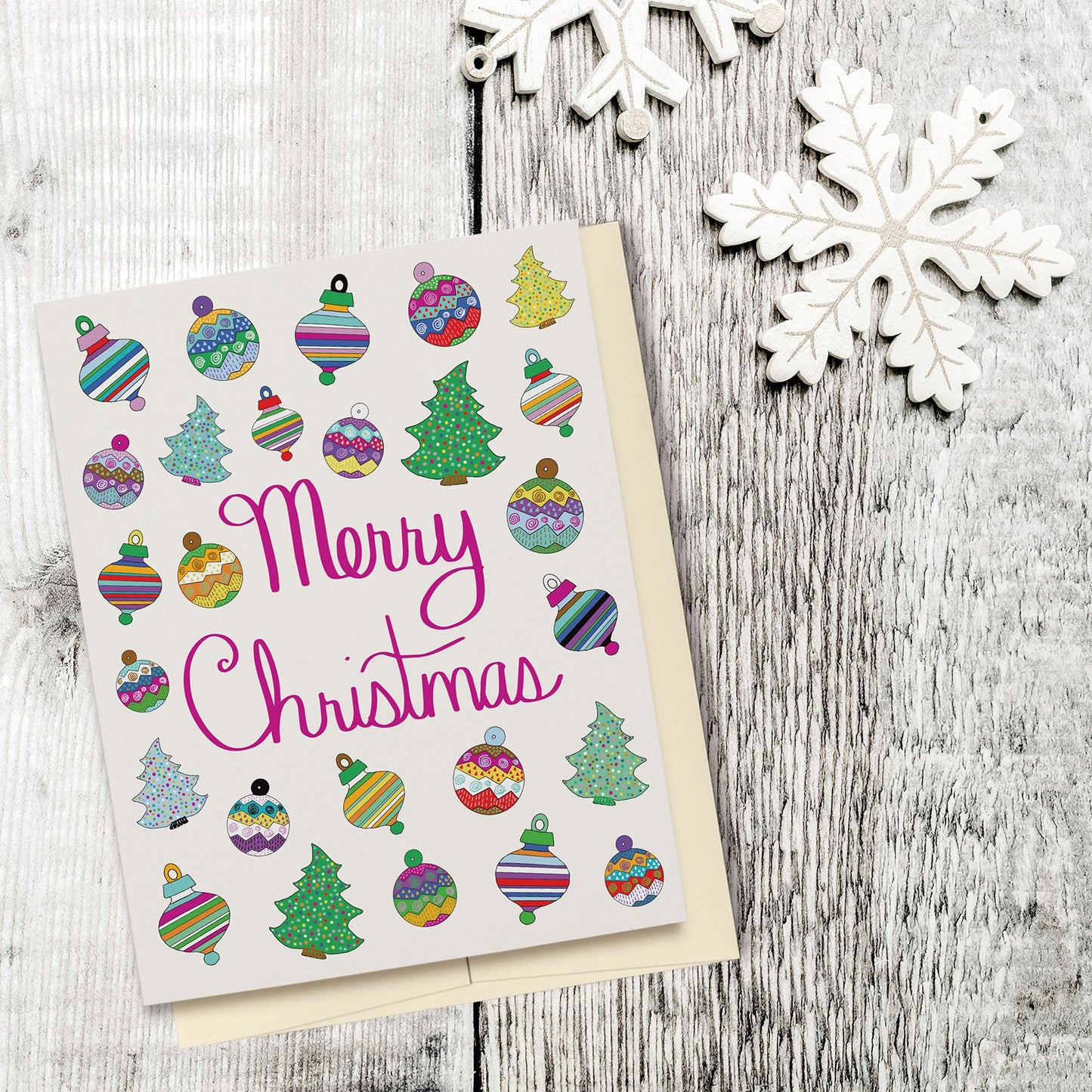 Fun & Festive Merry Christmas Card featuring hand drawn colorful ornaments & Christmas trees with fuchsia-colored hand lettered "Merry Christmas" script. Displayed on a wood background with wooden snowflake ornaments.