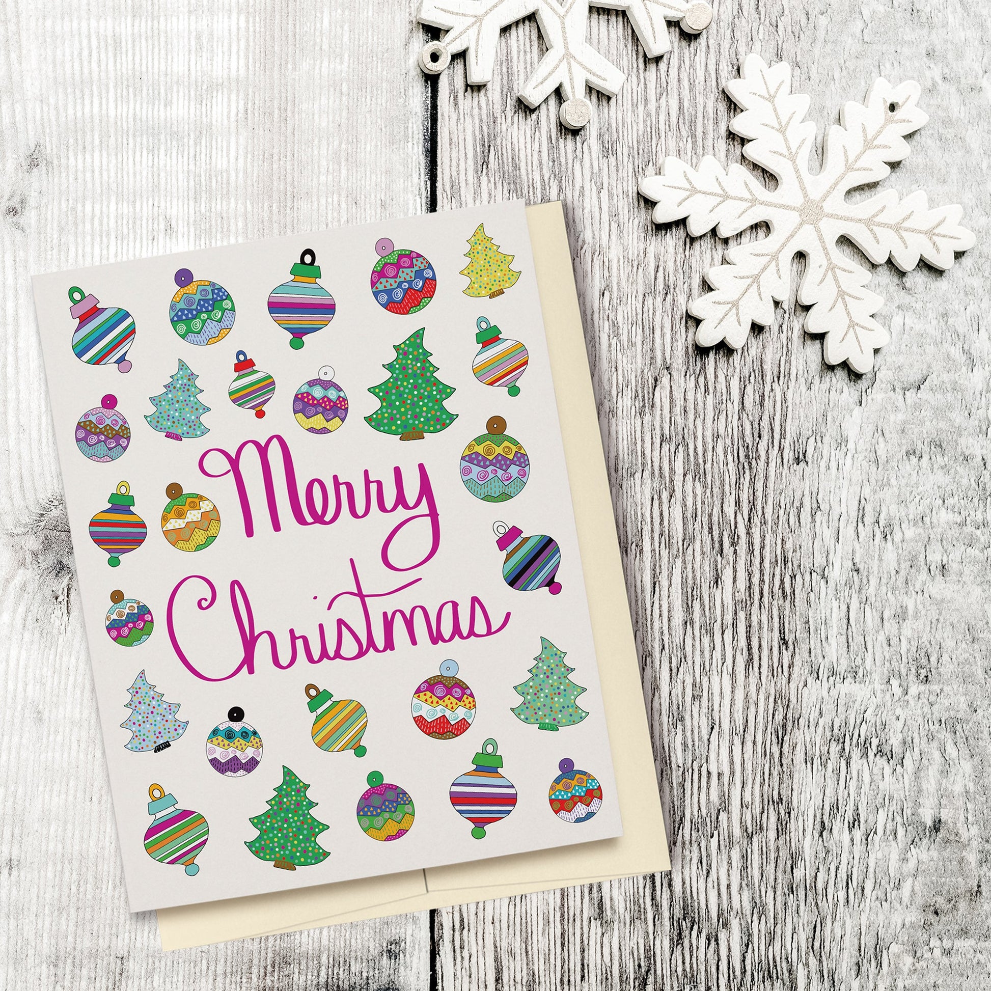 Fun & Festive Merry Christmas Card featuring hand drawn colorful ornaments & Christmas trees with fuchsia-colored hand lettered "Merry Christmas" script. Displayed on a wood background with wooden snowflake ornaments.