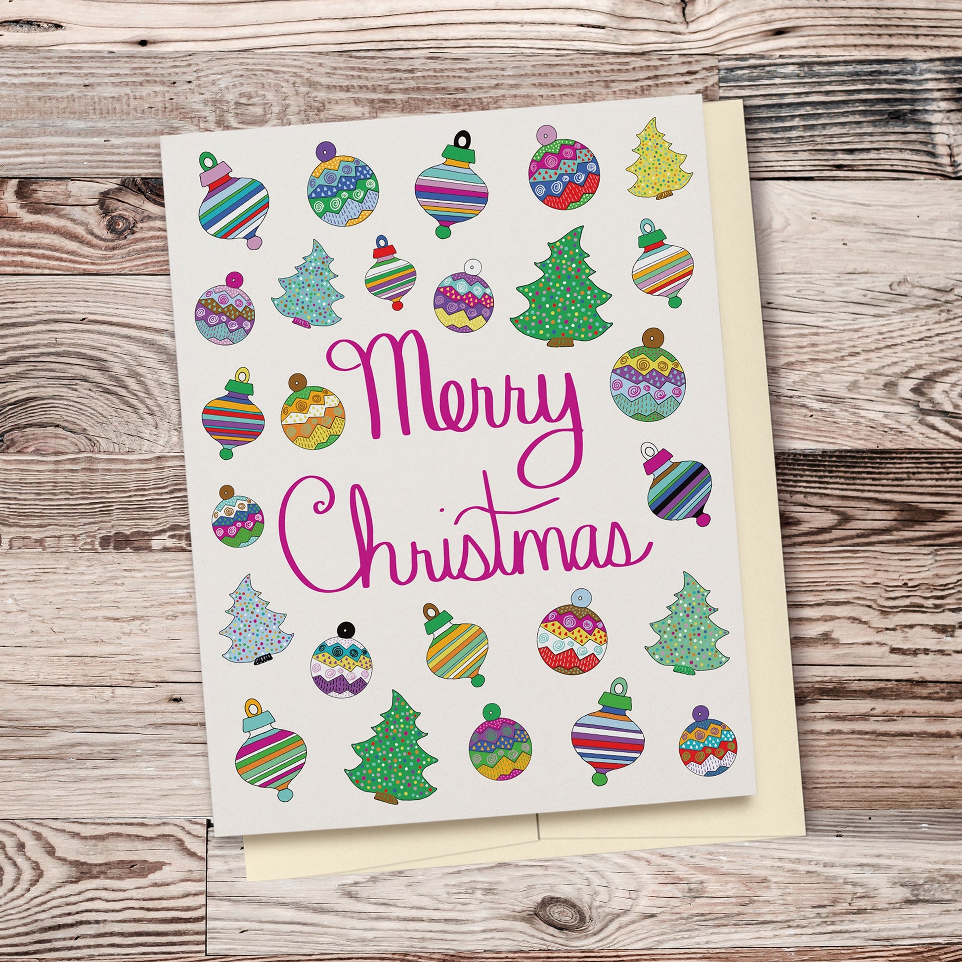 Fun & Festive Merry Christmas Card featuring hand drawn colorful ornaments & Christmas trees with fuchsia-colored hand lettered "Merry Christmas" script. Displayed on a wood background.
