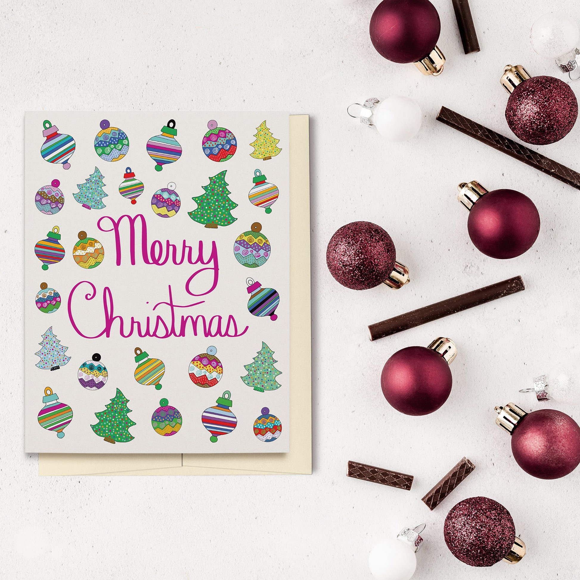 Fun & Festive Merry Christmas Card featuring hand drawn colorful ornaments & Christmas trees with fuchsia-colored hand lettered "Merry Christmas" script. Displayed on a white cement backgrounds with red ornaments and chocolate candy.