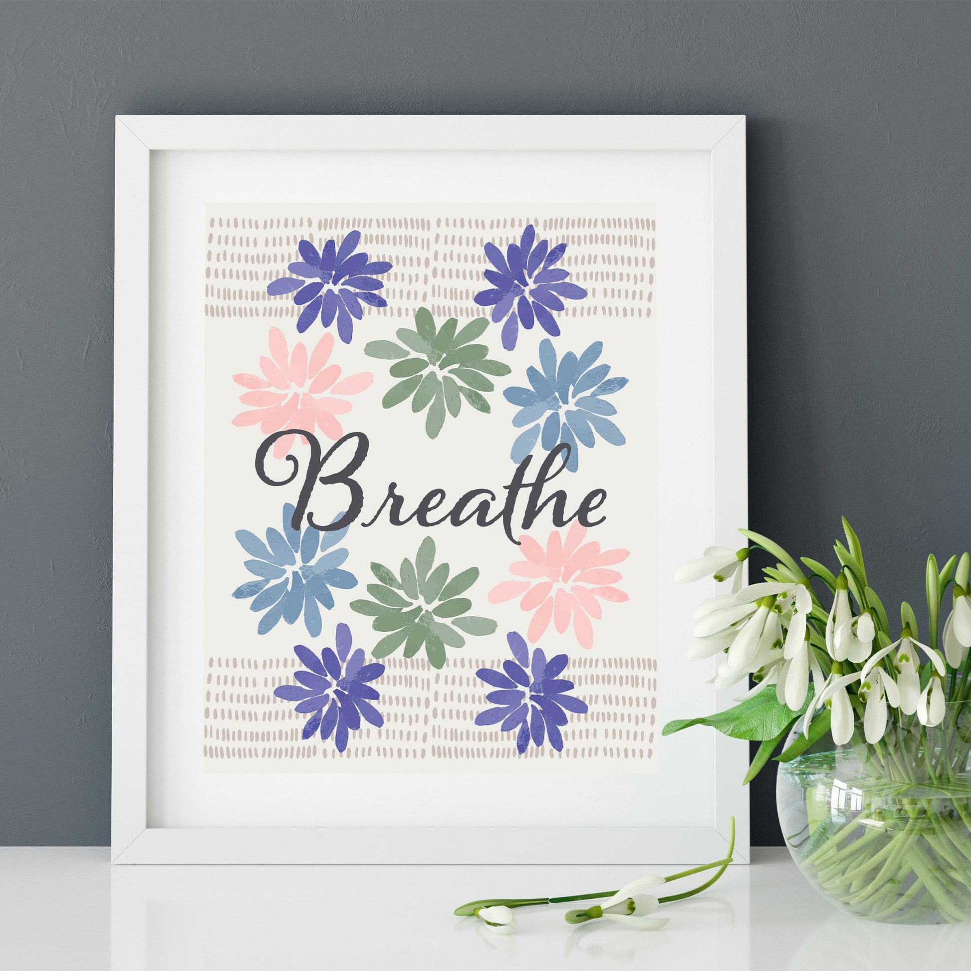 An eco-friendly art print featuring gouache painted flowers and marks with the word "Breathe" in script, shown in a white frame on a white table with white flowers in a vase against a gray wall.