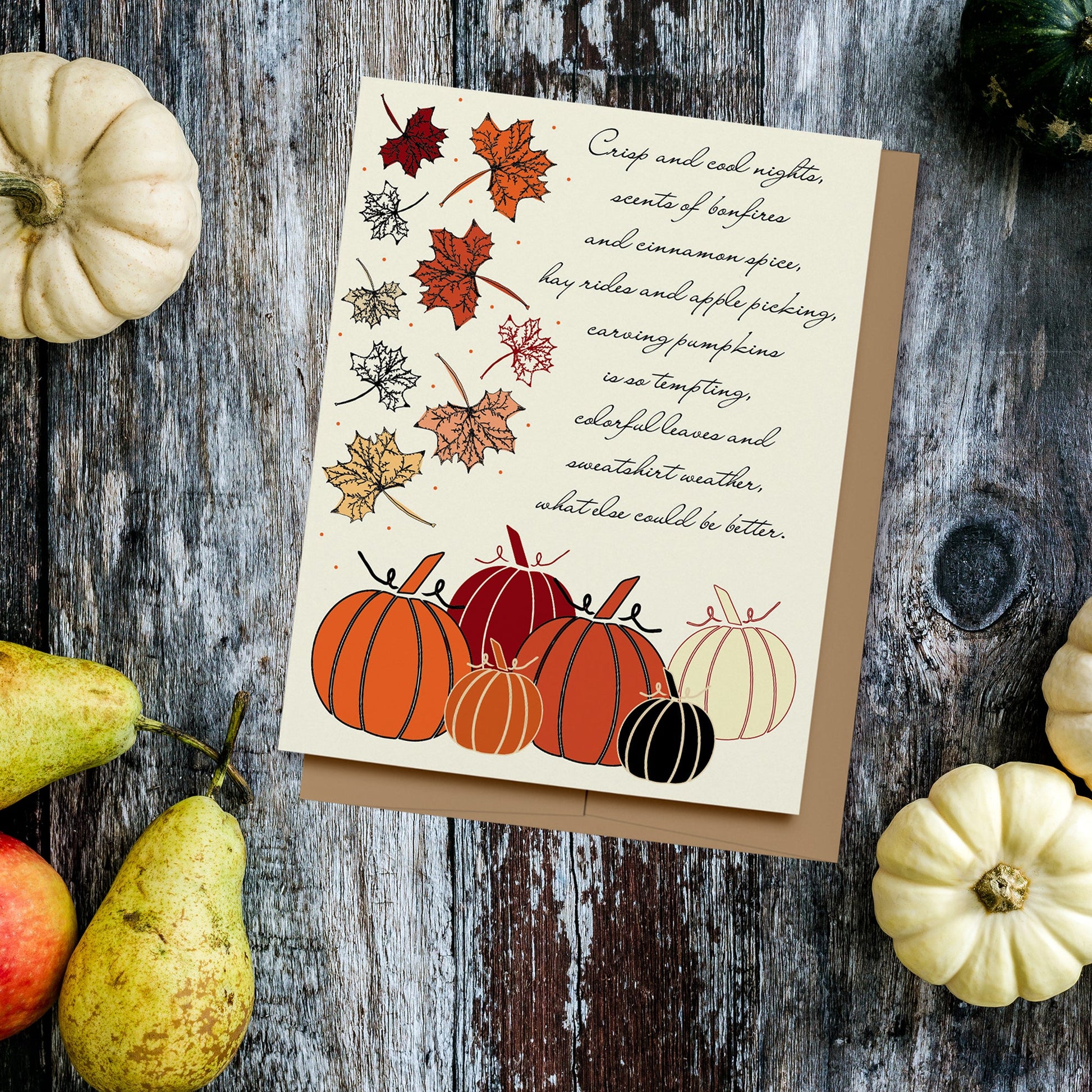 A fun card to celebrate the autumn season featuring hand drawn pumpkins and autumn leaves on a cream background which reads, "Crisp and cool nights, scents of bonfires and cinnamon spice, hay rides and apple picking, carving pumpkins is so tempting, colorful leaves and sweatshirt weather, what else could be better." Displayed on a wood background surrounded by gourds, pears and apple.