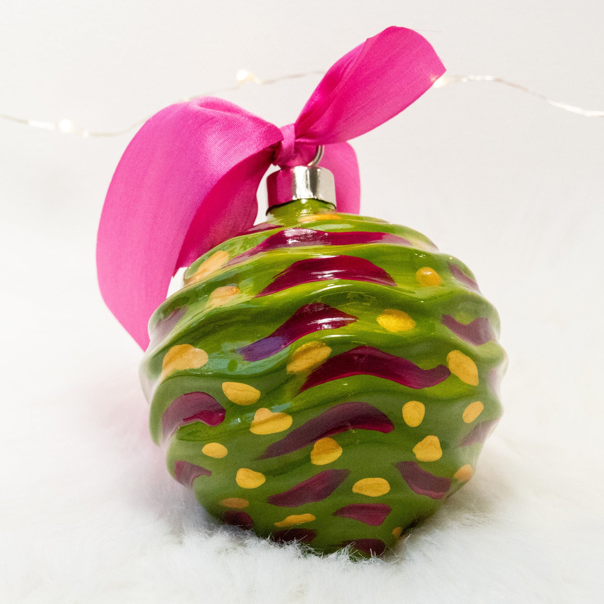 The Avery Hand Painted Ornament features a green gold base coat with rose violet and gold marks. Painted using fluid acrylic and acryla gouache paints. Displayed on white faux fur with fairy lights in the background.