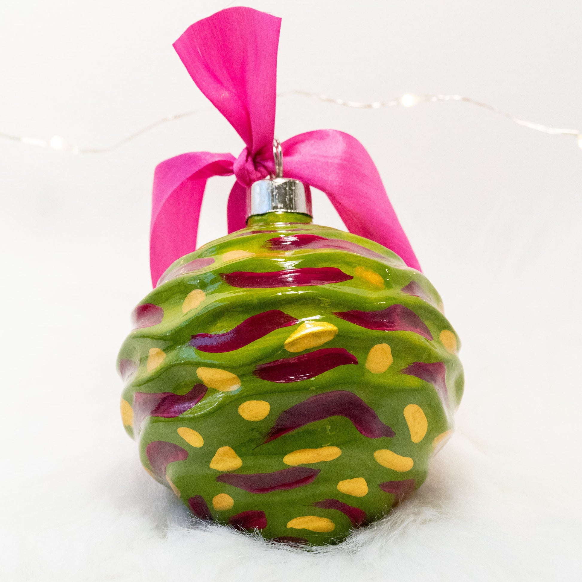 The Avery Hand Painted Ornament features a green gold base coat with rose violet and gold marks. Painted using fluid acrylic and acryla gouache paints. Displayed on white faux fur with fairy lights in the background.