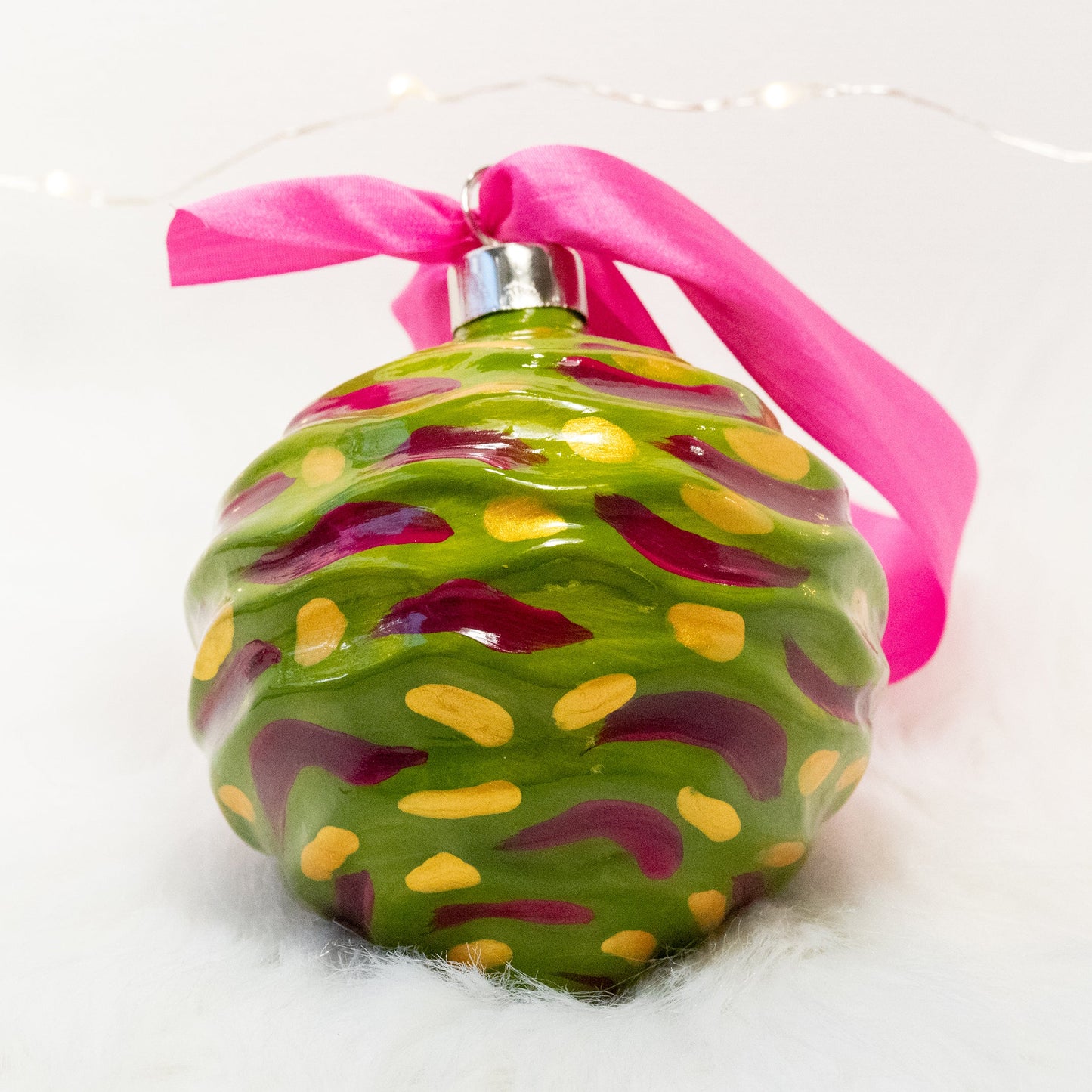The Avery Hand Painted Ornament features a green gold base coat with rose violet and gold marks. Painted using fluid acrylic and acryla gouache paints. Displayed on white faux fur with fairy lights in the background.