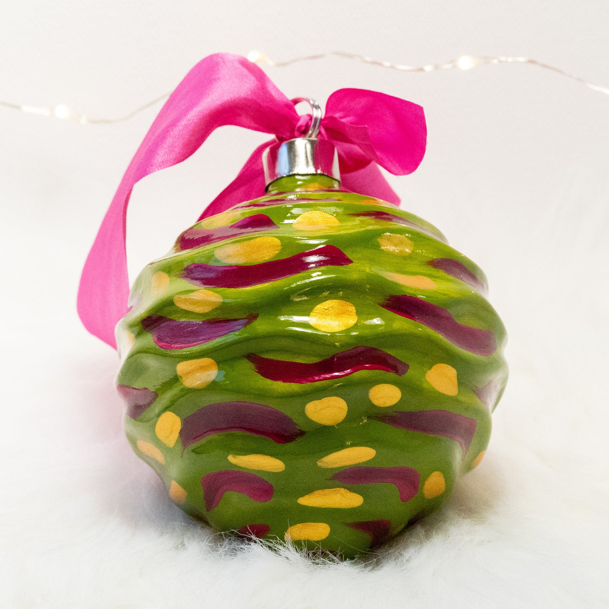 The Avery Hand Painted Ornament features a green gold base coat with rose violet and gold marks. Painted using fluid acrylic and acryla gouache paints. Displayed on white faux fur with fairy lights in the background.