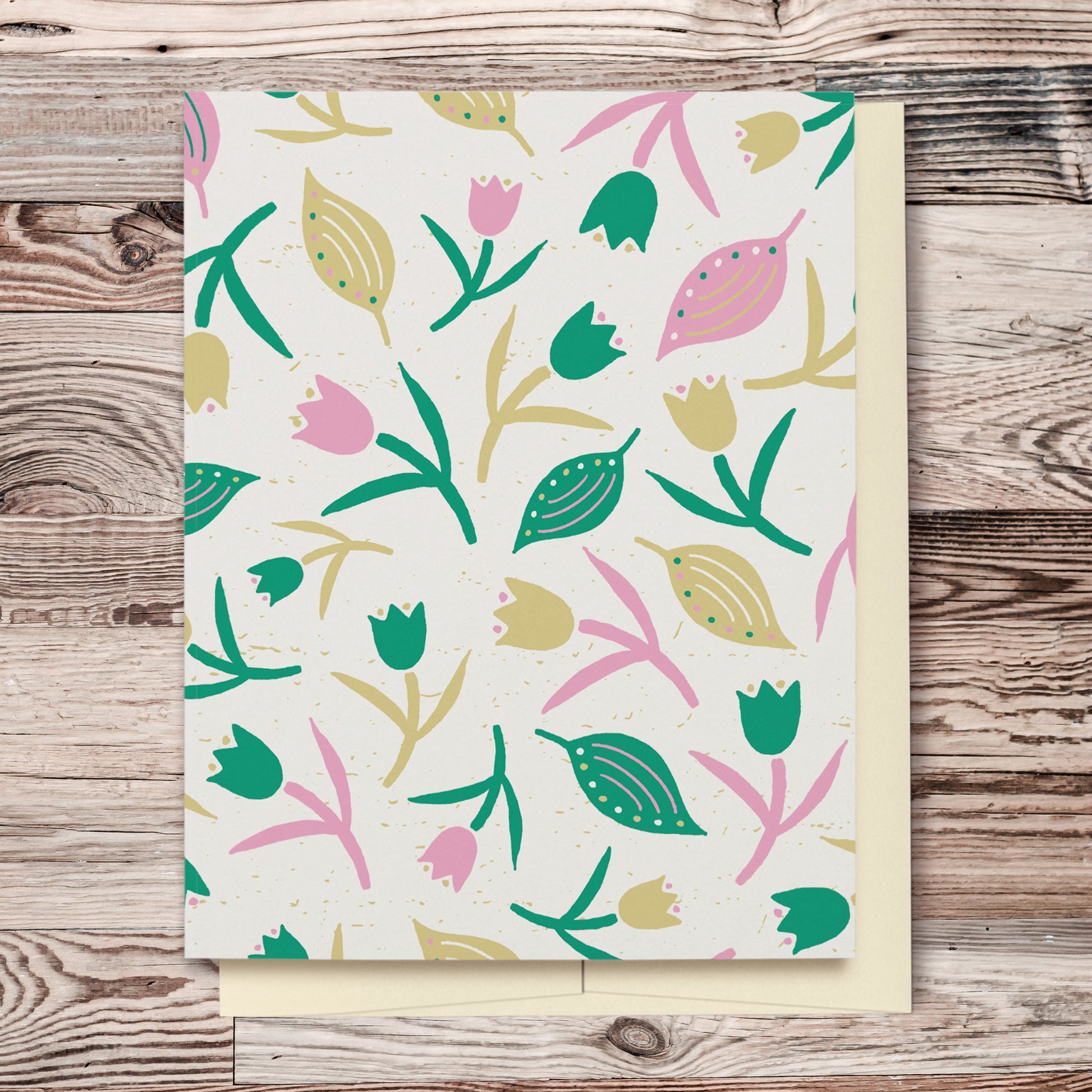 Tulips & Leaves Cream, Green & Pink Blank Card features a pattern with tulips and leaves in shades of green and pink on a cream background. The pattern extends over the back of the card too. Displayed on a wooden background.