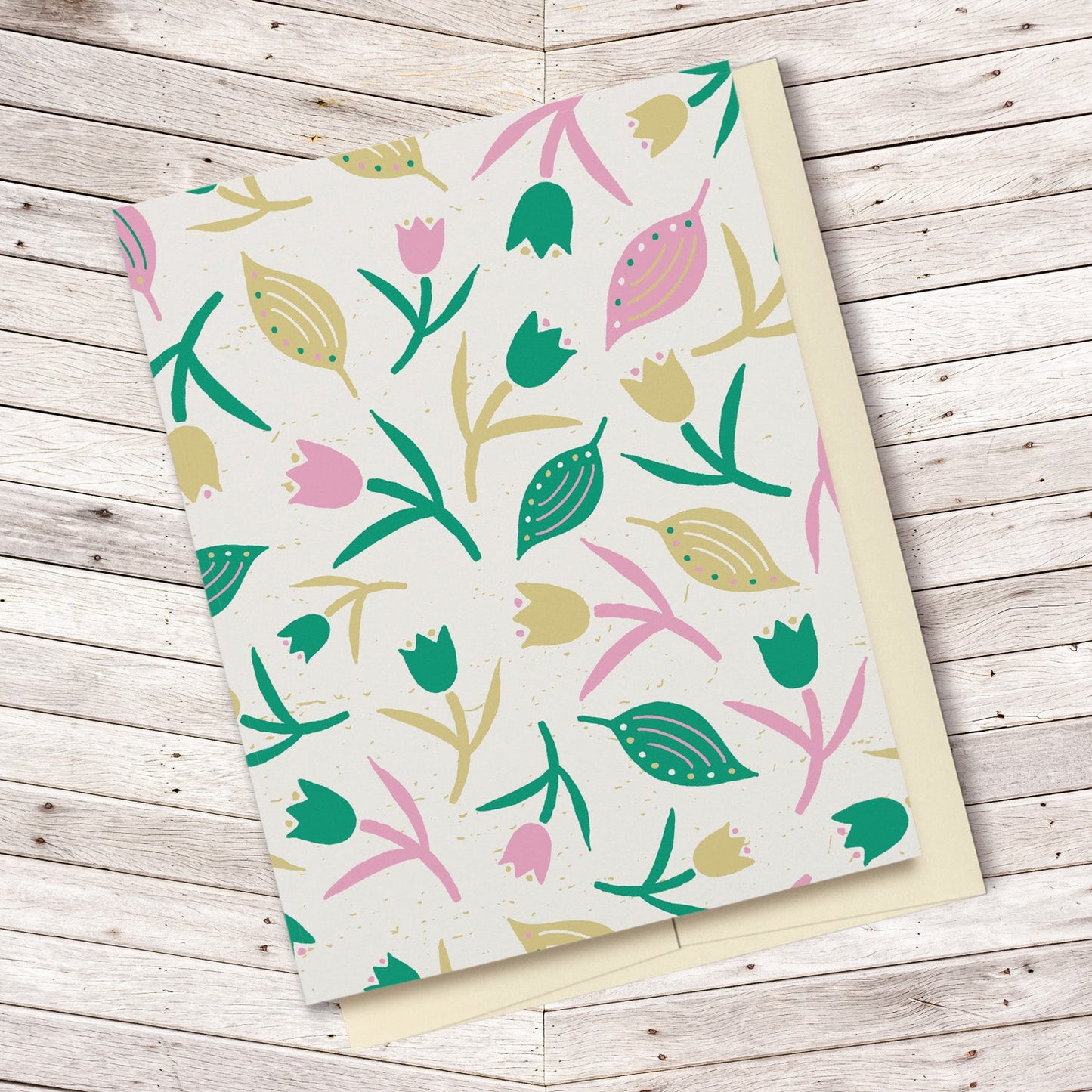 Tulips & Leaves Cream, Green & Pink Blank Card features a pattern with tulips and leaves in shades of green and pink on a cream background. The pattern extends over the back of the card too. Displayed on a wooden background.