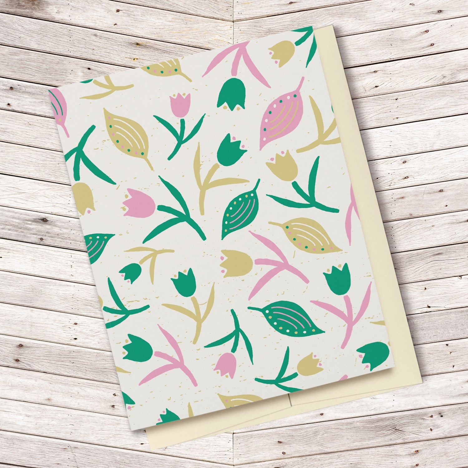 Tulips & Leaves Cream, Green & Pink Blank Card features a pattern with tulips and leaves in shades of green and pink on a cream background. The pattern extends over the back of the card too. Displayed on a wooden background.