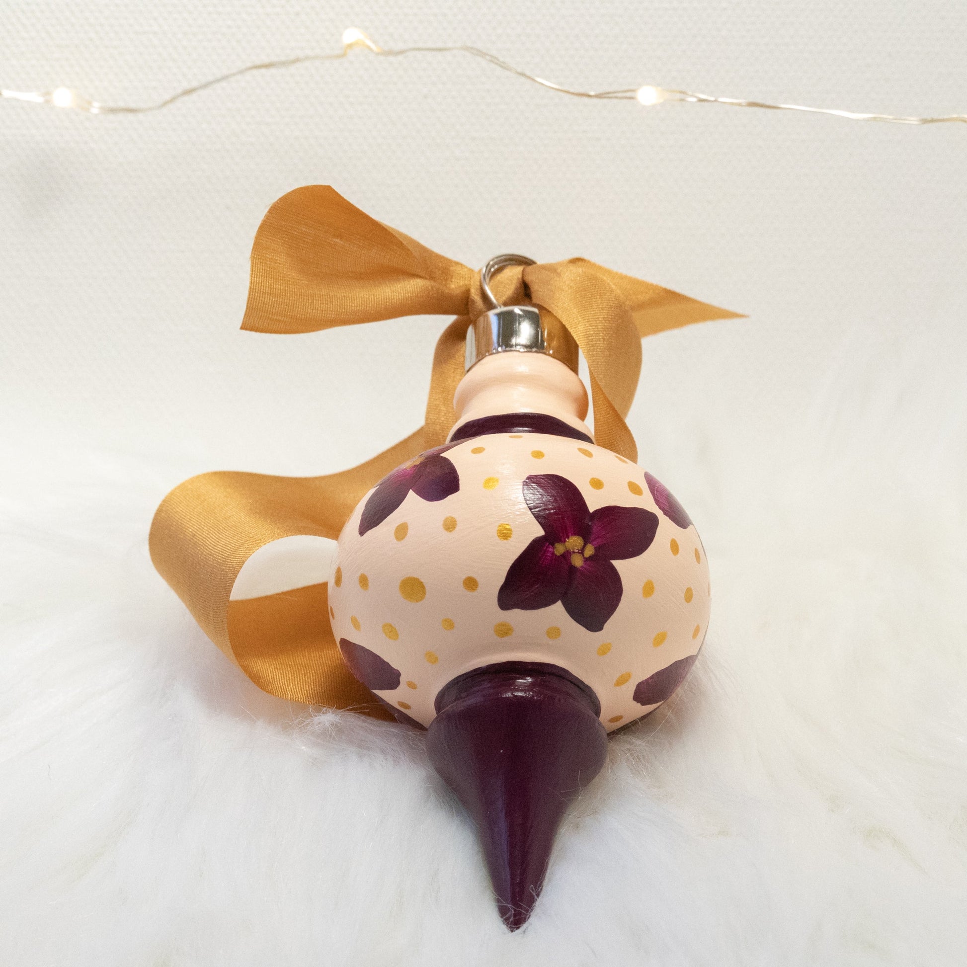The Charlotte Hand Painted Ornament features a light peach and deep magenta base coat, deep magenta flowers with gold polka dots. Painted using fluid acrylic and acryla gouache paints. Displayed on white faux fur with fairy lights in the background.