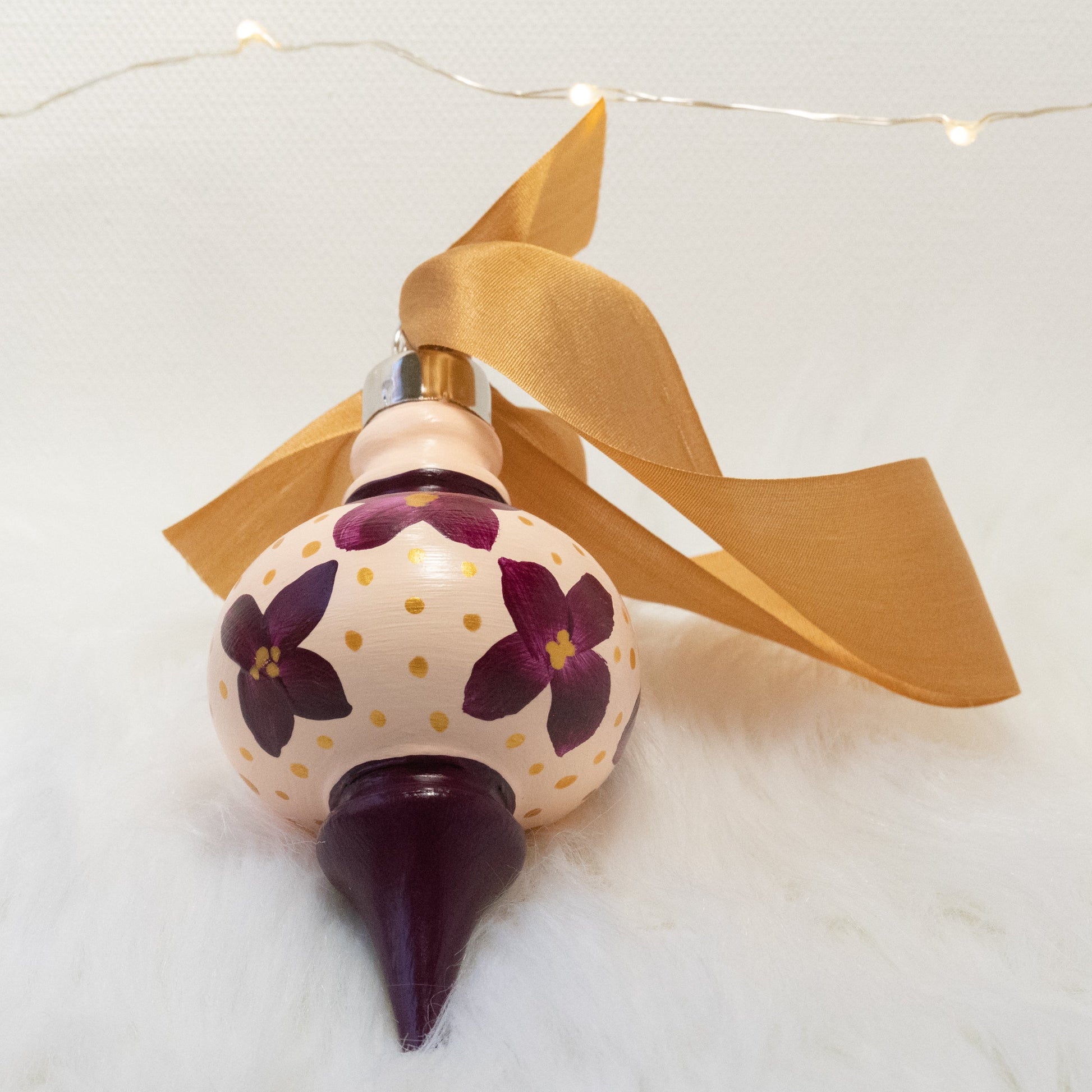 The Charlotte Hand Painted Ornament features a light peach and deep magenta base coat, deep magenta flowers with gold polka dots. Painted using fluid acrylic and acryla gouache paints. Displayed on white faux fur with fairy lights in the background.