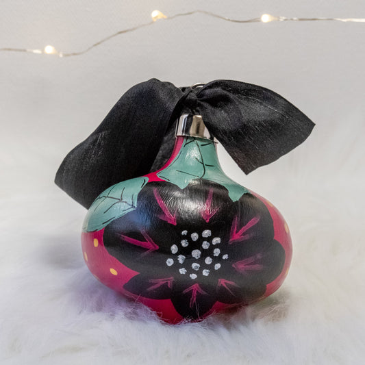 The Elizabeth Hand Painted Ornament features a rose violet base coat, black flowers with sage green leaves and black, rose violet, gold & metallic white accent details. Painted using acryla gouache paints. Displayed on white faux fur with fairy lights in the background.