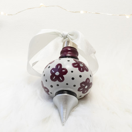 The Helena Hand Painted Ornament features a light gray, silver and deep magenta base coat, deep magenta flowers with black and silver accents. Painted using fluid acrylic and acryla gouache paints. Displayed on white faux fur with fairy lights in the background.