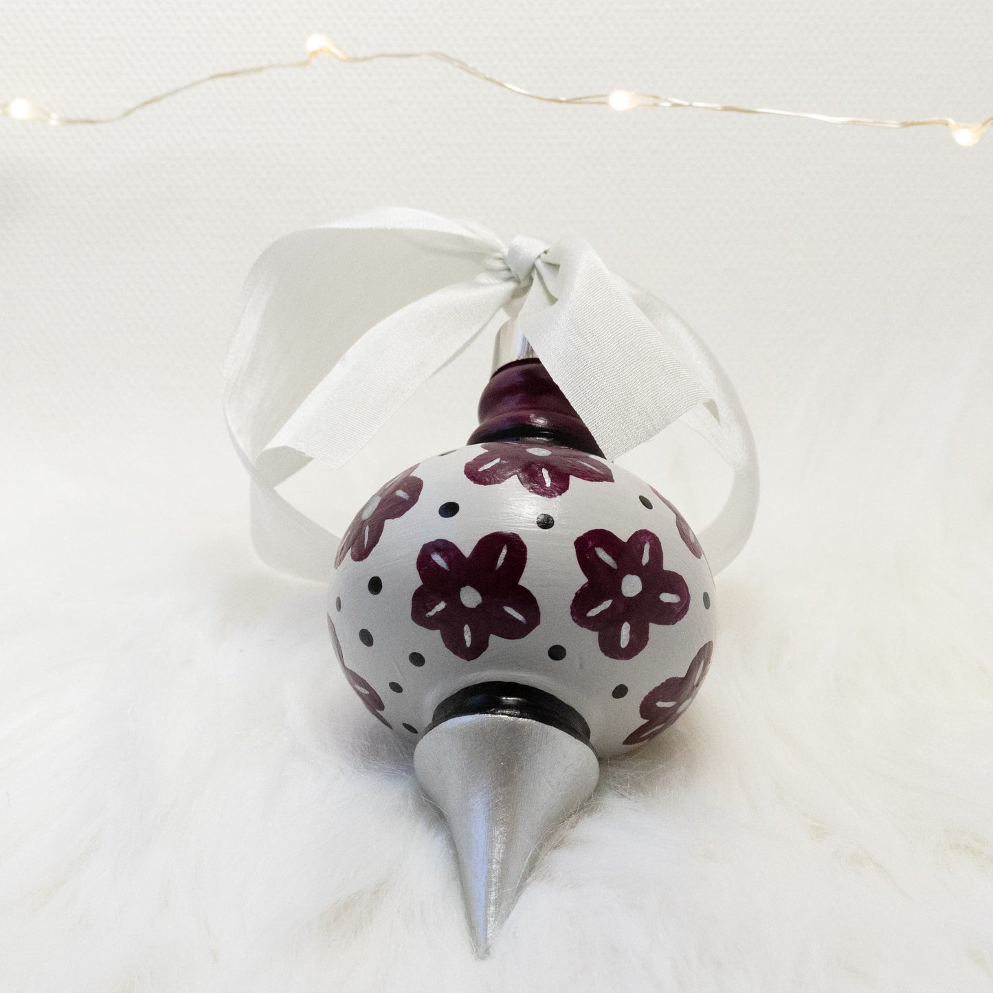 The Helena Hand Painted Ornament features a light gray, silver and deep magenta base coat, deep magenta flowers with black and silver accents. Painted using fluid acrylic and acryla gouache paints. Displayed on white faux fur with fairy lights in the background.