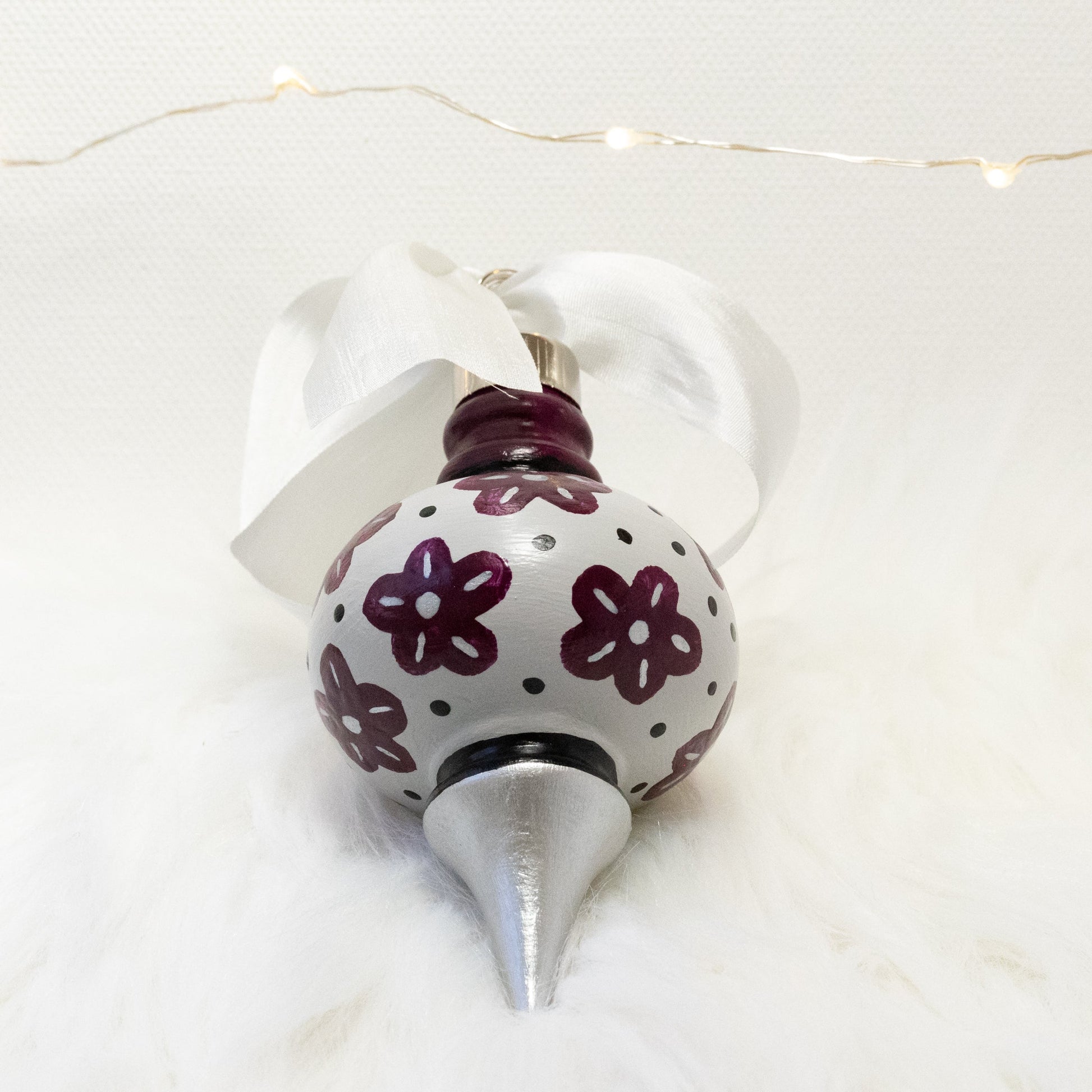 The Helena Hand Painted Ornament features a light gray, silver and deep magenta base coat, deep magenta flowers with black and silver accents. Painted using fluid acrylic and acryla gouache paints. Displayed on white faux fur with fairy lights in the background.