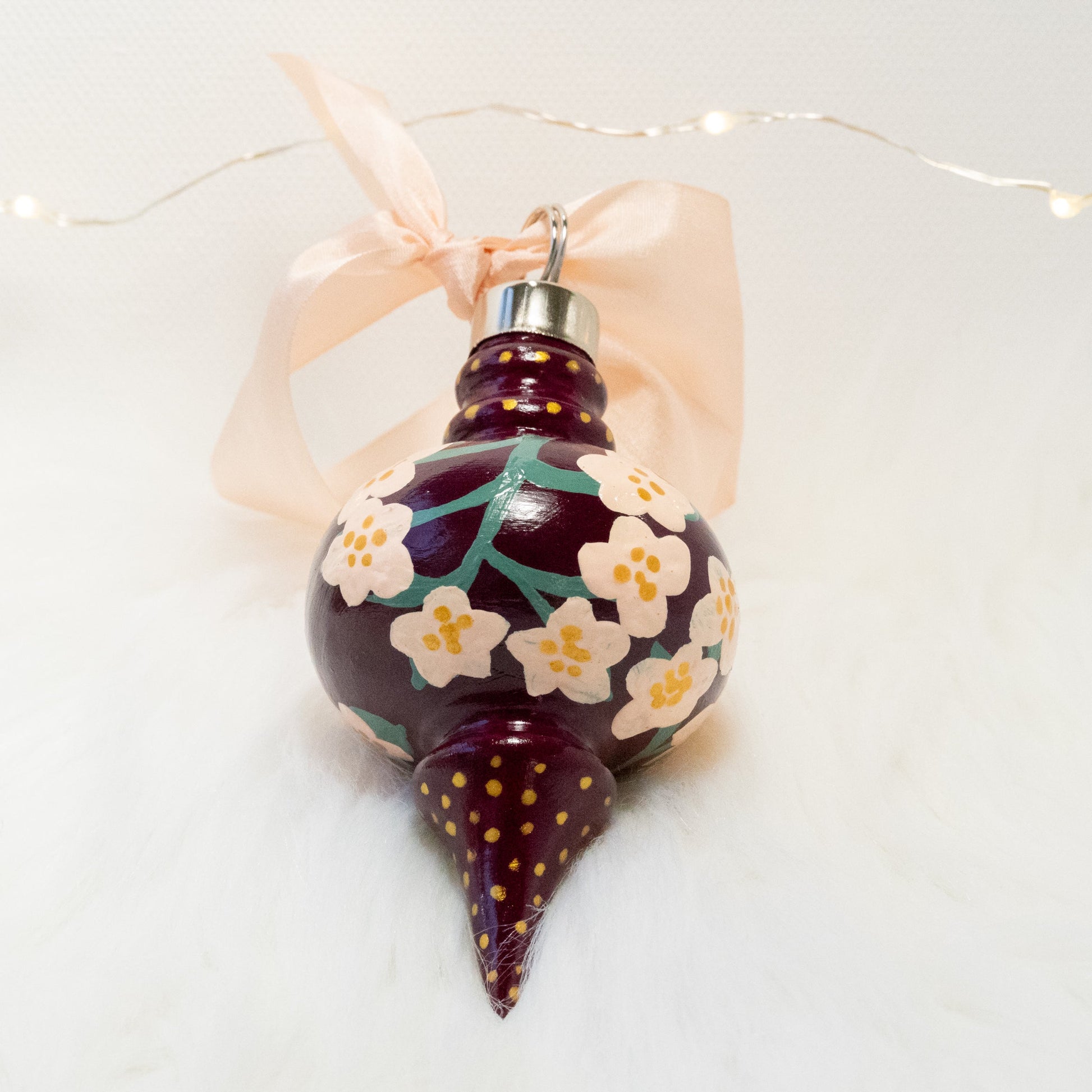The Holly Hand Painted Ornament features a deep magenta base coat, light peach flowers with sage green stems and gold polka dots. Painted using fluid acrylic and acryla gouache paints. Displayed on white faux fur with fairy lights in the background.
