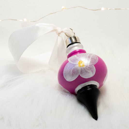 The Kiki Hand Painted Ornament features a fuchsia and black base coat, metallic white flowers with metallic white, gold and black accents. Painted using fluid acrylic and acryla gouache paints. Displayed on white faux fur with fairy lights in the background.