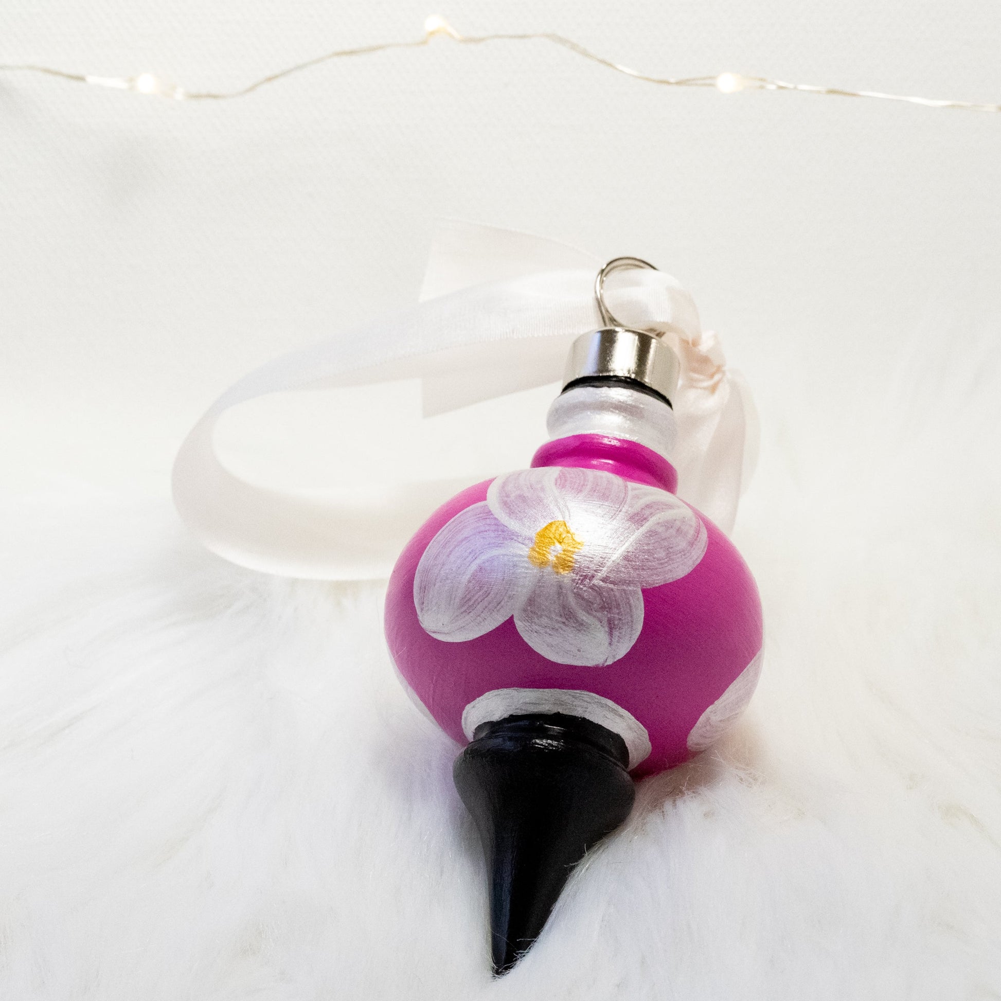 The Kiki Hand Painted Ornament features a fuchsia and black base coat, metallic white flowers with metallic white, gold and black accents. Painted using fluid acrylic and acryla gouache paints. Displayed on white faux fur with fairy lights in the background.