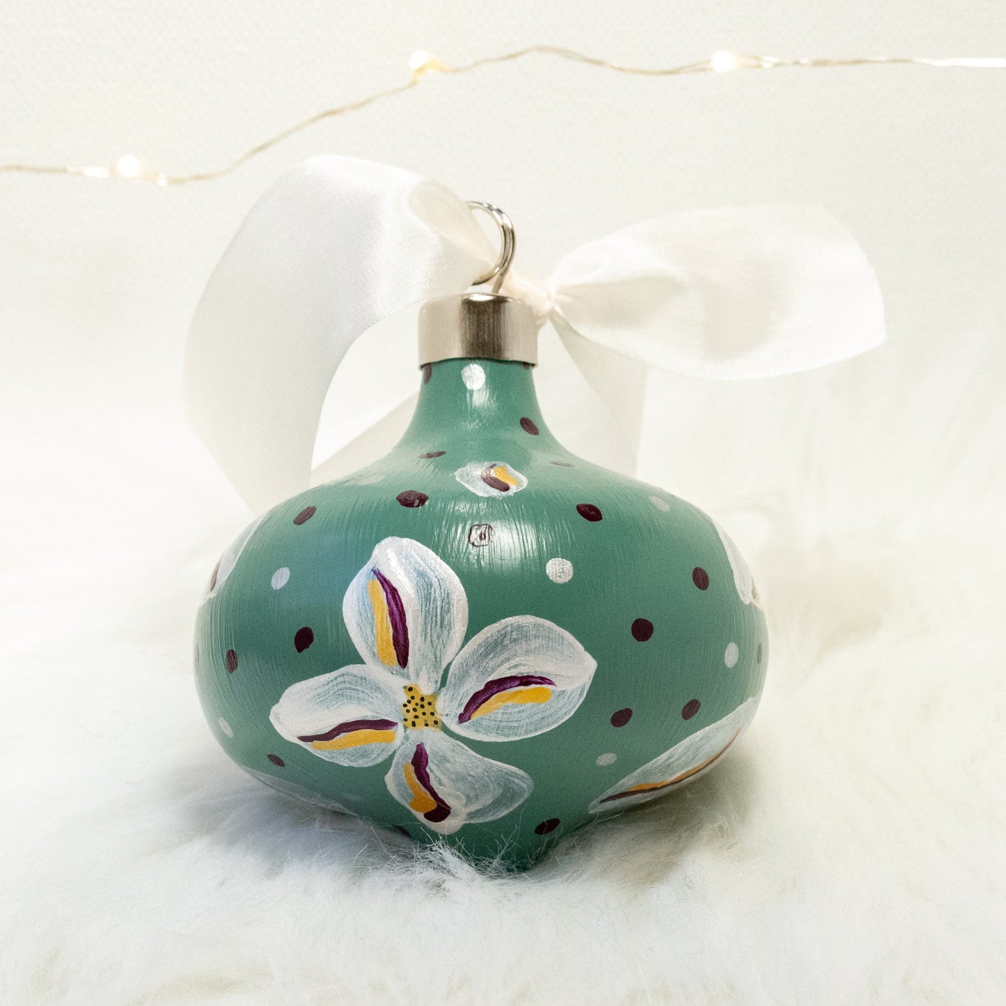 The Kristina Hand Painted Ornament features a sage green base coat, metallic white flowers with deep magenta and gold accent details. Painted using acryla gouache paints. Displayed on white faux fur with fairy lights in the background.