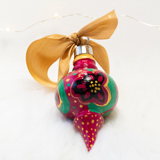 The Lois Hand Painted Ornament features a rose violet base coat, black flowers with gold and green polka dots and accents. Painted using fluid acrylic and acryla gouache paints. Displayed on white faux fur with fairy lights in the background.