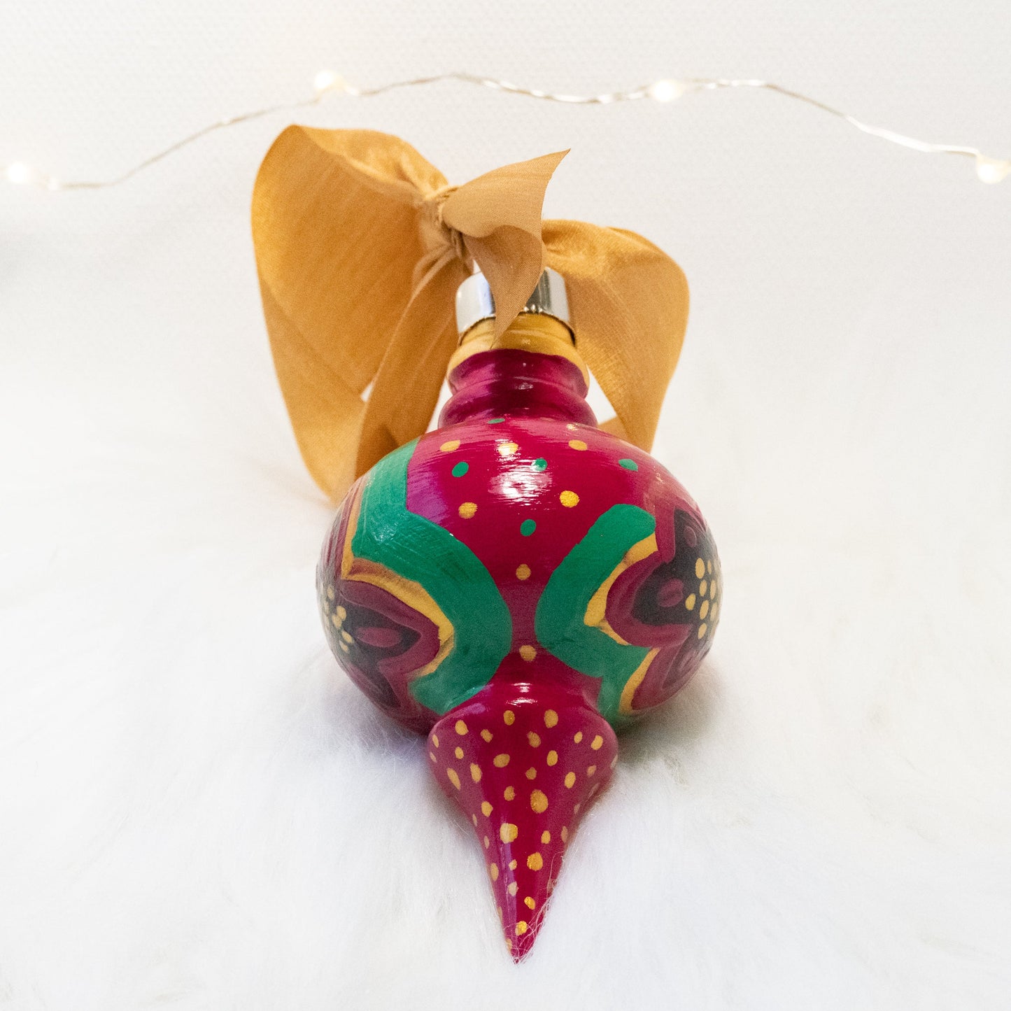 The Lois Hand Painted Ornament features a rose violet base coat, black flowers with gold and green polka dots and accents. Painted using fluid acrylic and acryla gouache paints. Displayed on white faux fur with fairy lights in the background.