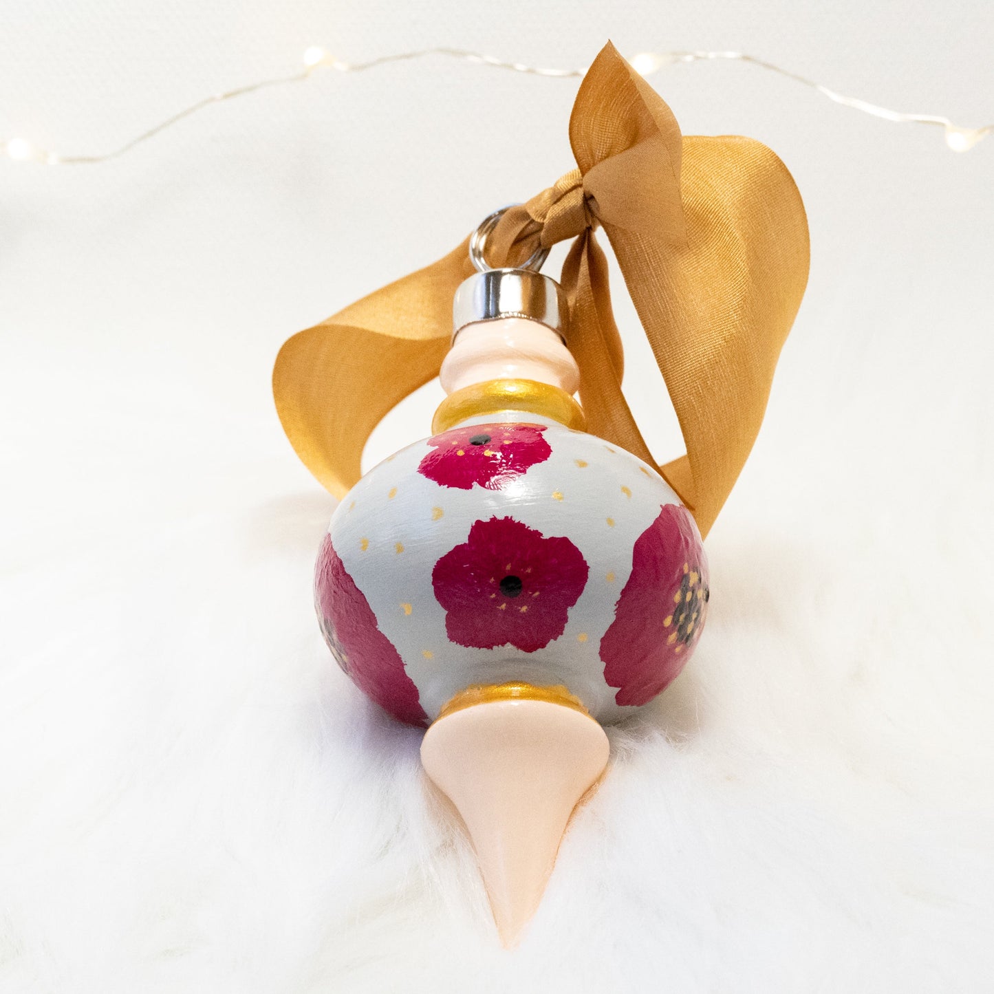 The Lulu Hand Painted Ornament features a light gray and peach base coat, rose violet flowers with gold polka dots and black and gold accents. Painted using fluid acrylic and acryla gouache paints. Displayed on white faux fur with fairy lights in the background.
