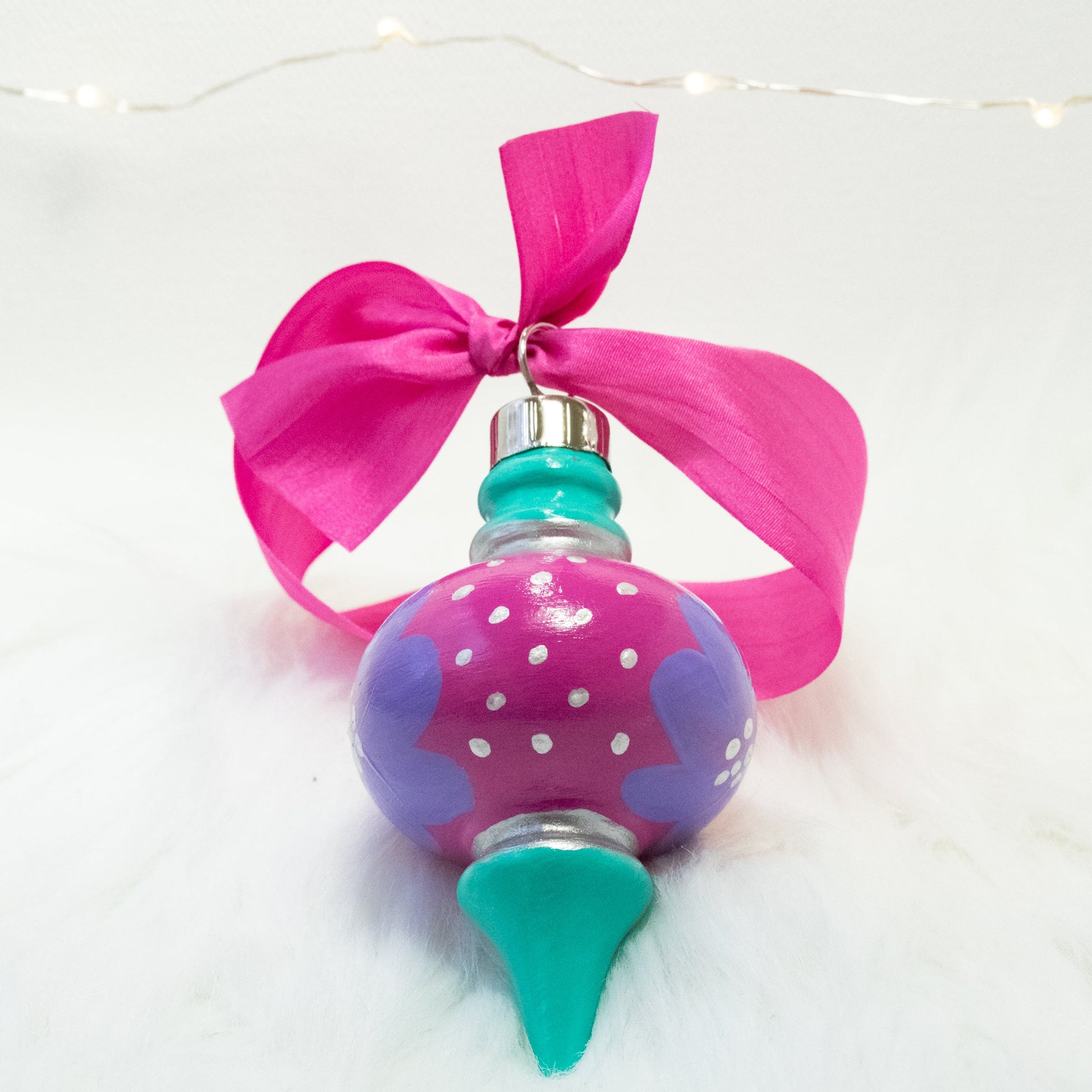 The Maxie Hand Painted Ornament features a fuchsia and bright aqua green base coat, lilac purple flowers with silver accents. Painted using fluid acrylic and acryla gouache paints. Displayed on white faux fur with fairy lights in the background.